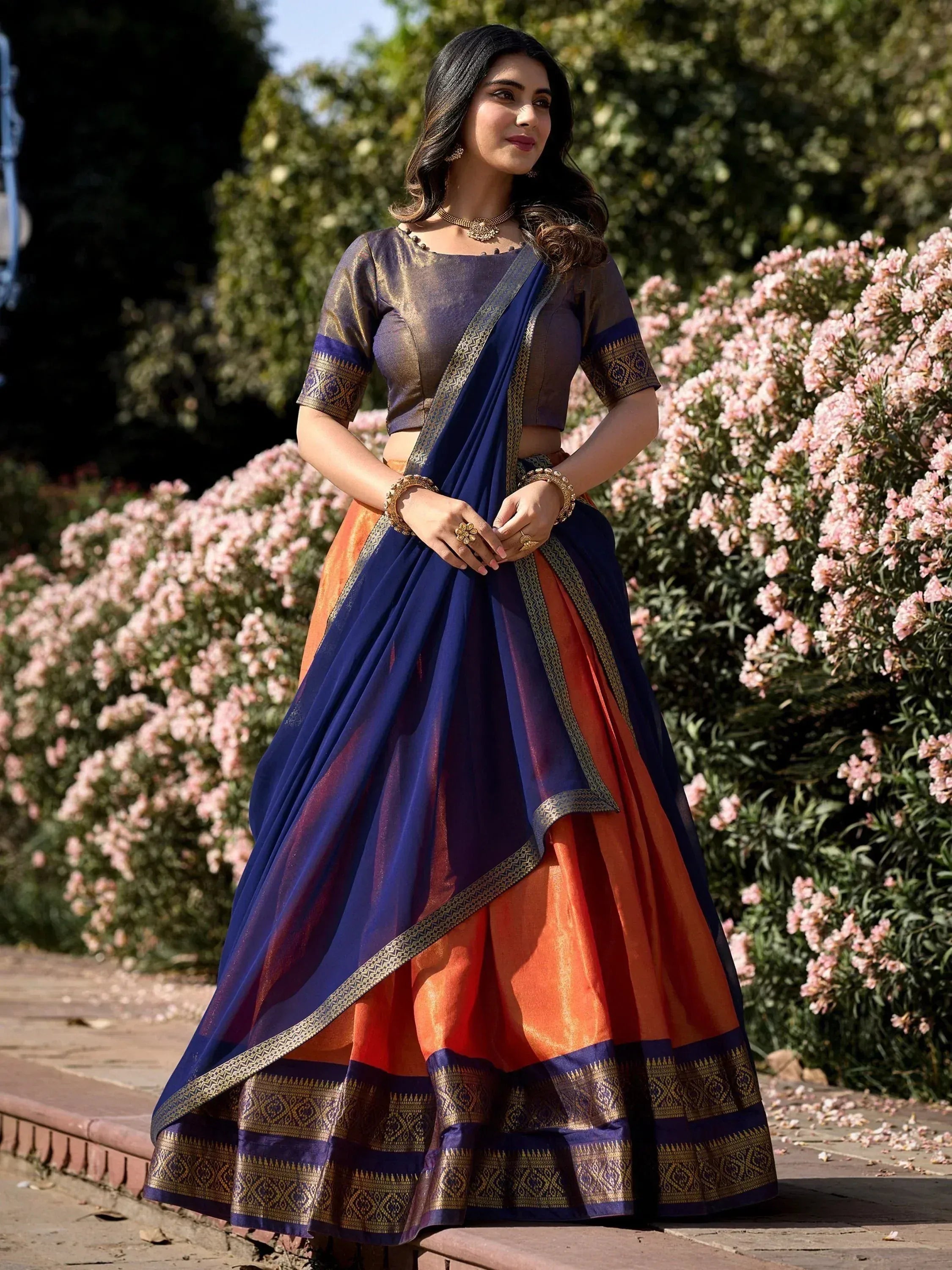 Woman in blue and orange designer lehenga choli with gold embroidery, standing in garden