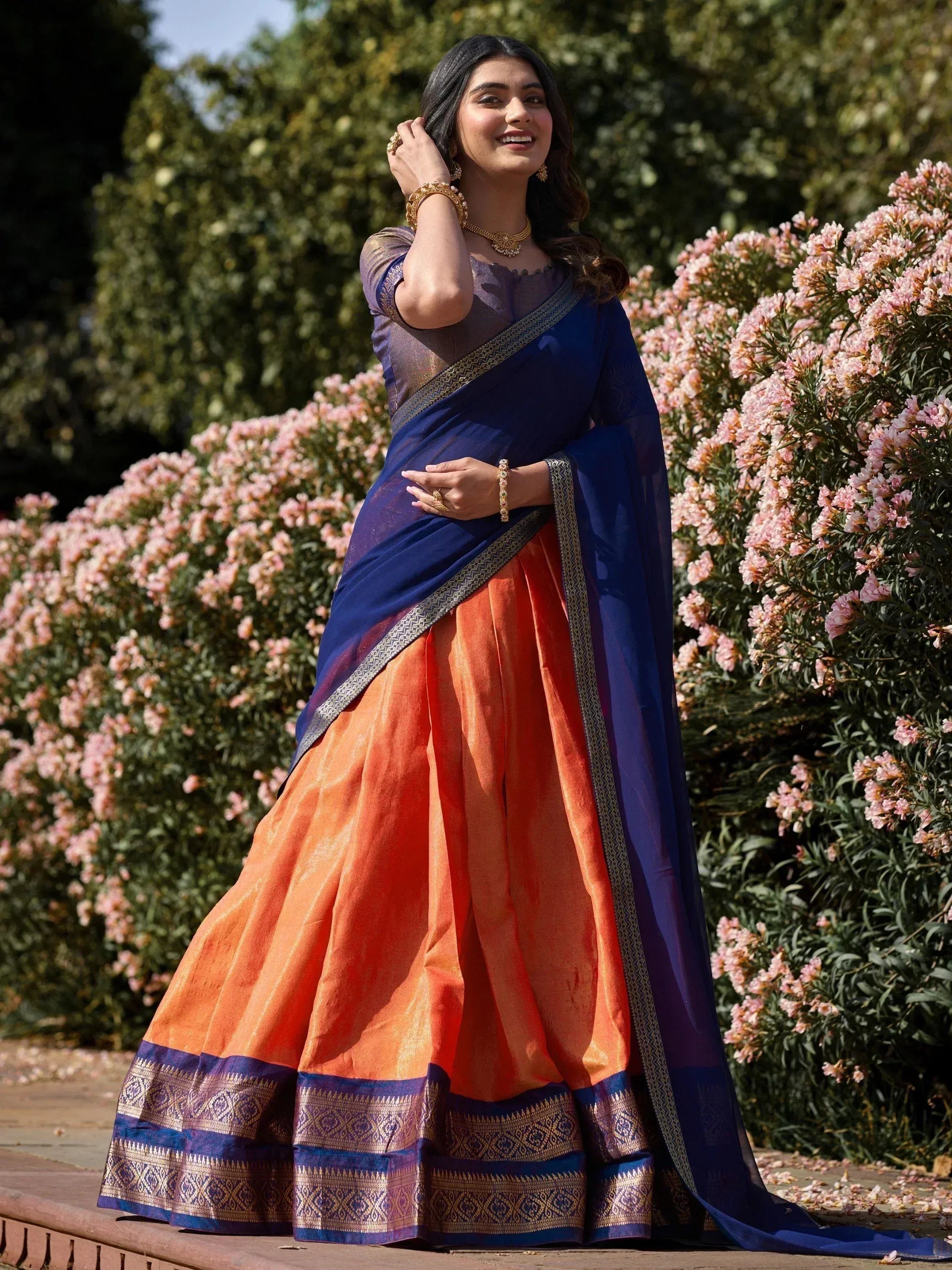 Woman in vibrant orange and navy blue saree with gold border, styled with traditional jewelry in garden setting