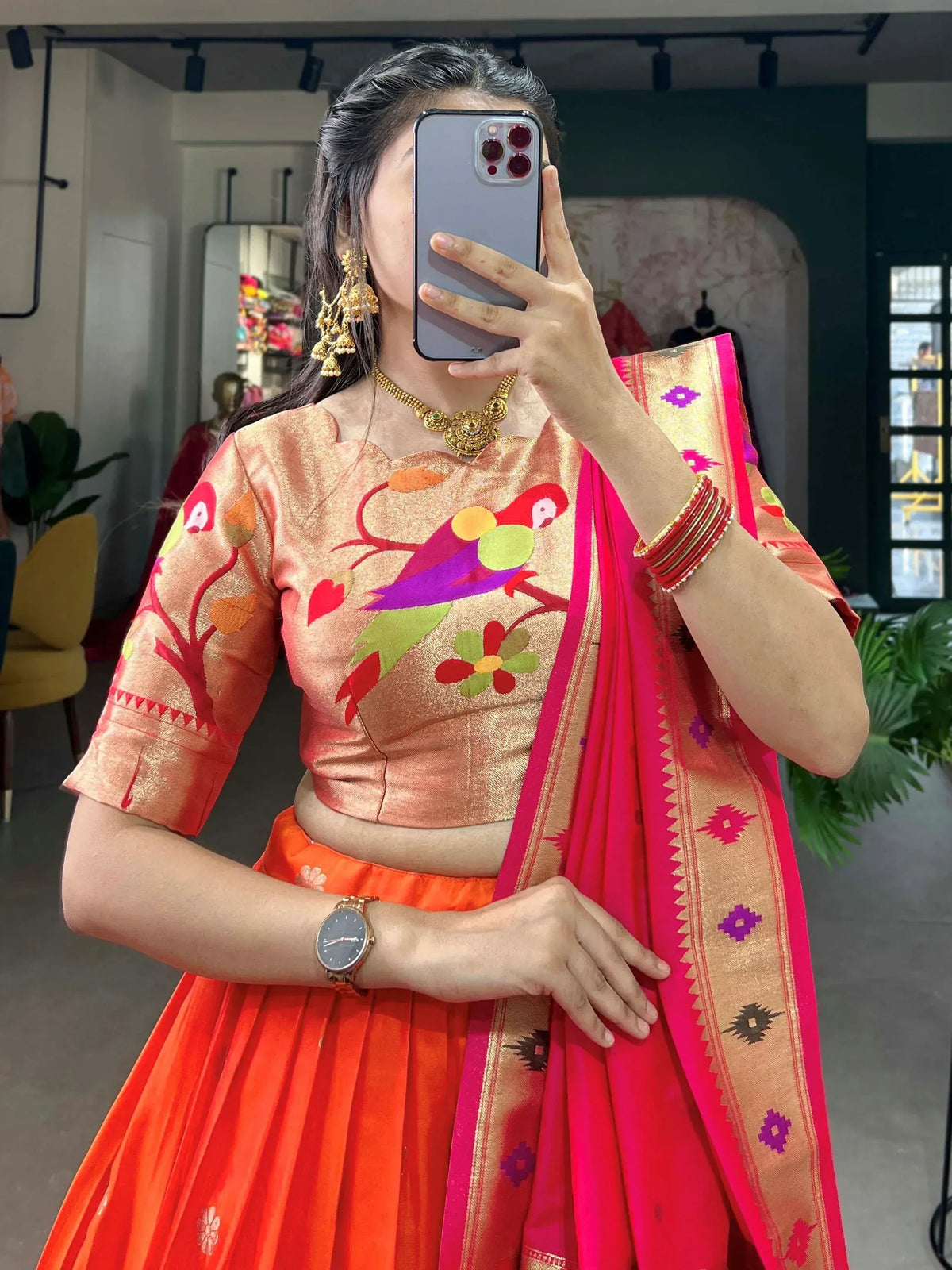 Woman in vibrant orange lehenga choli with parrot embroidery, paired with pink saree dupatta, gold jewelry