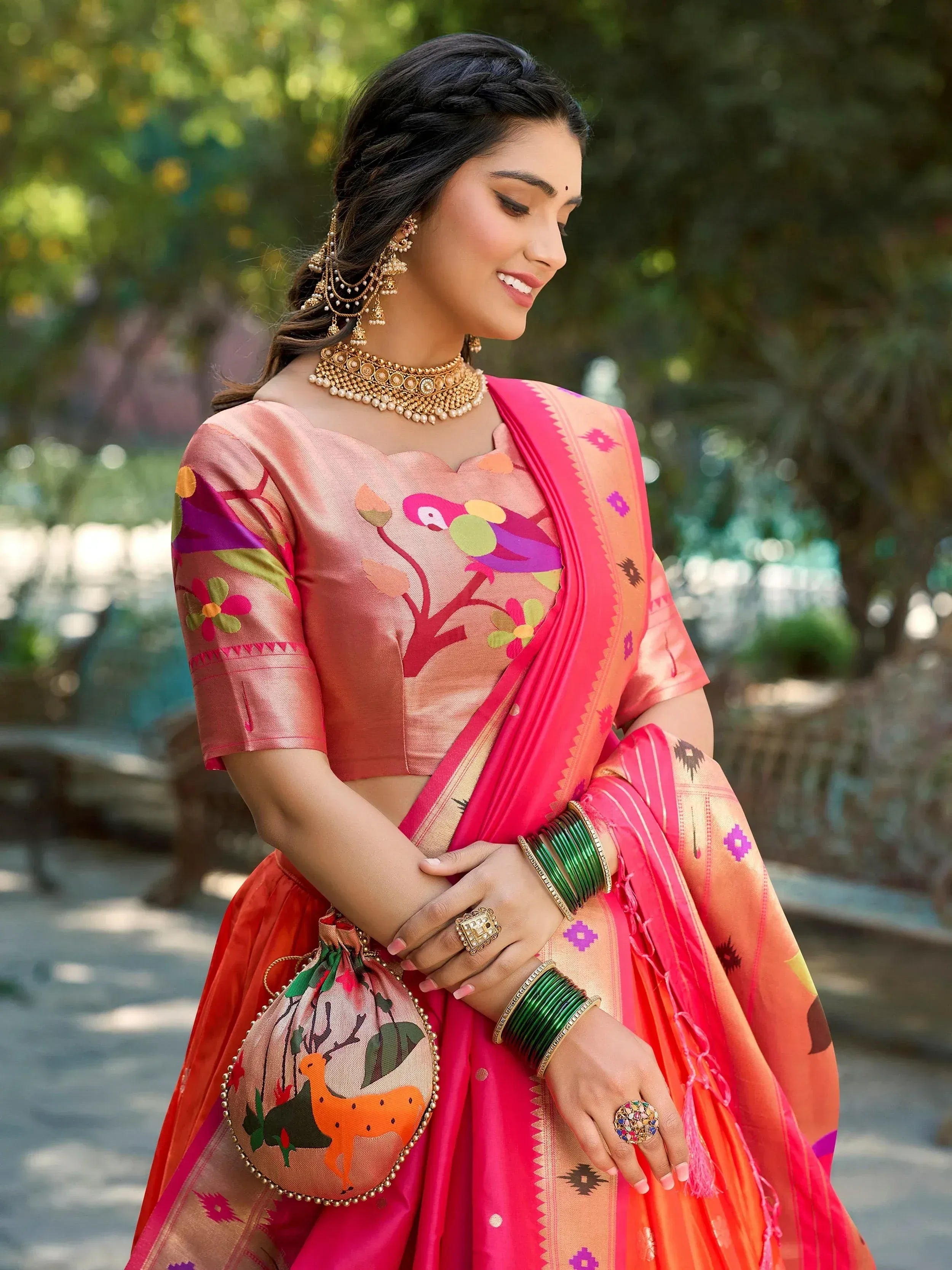 Woman in vibrant pink and orange printed lehenga choli with floral and bird motifs, paired with traditional gold jewelry and green bangles.