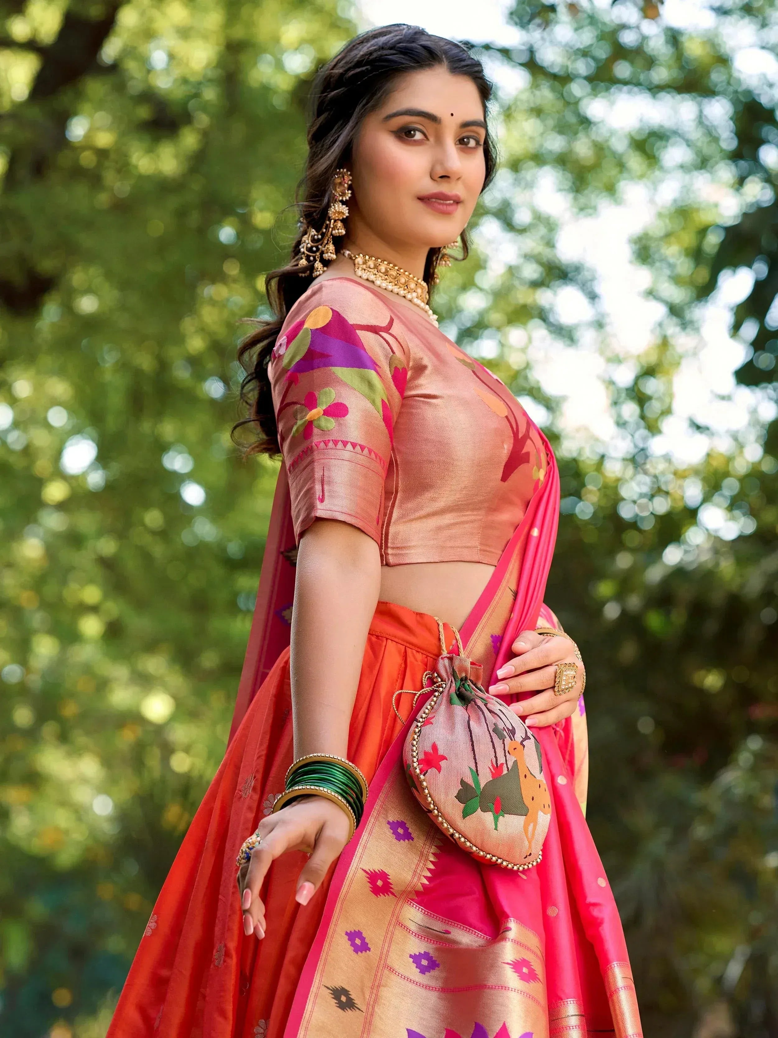 Woman in vibrant pink and orange embroidered lehenga choli with floral blouse, traditional jewelry, and matching potli bag, styled for festive occasions.