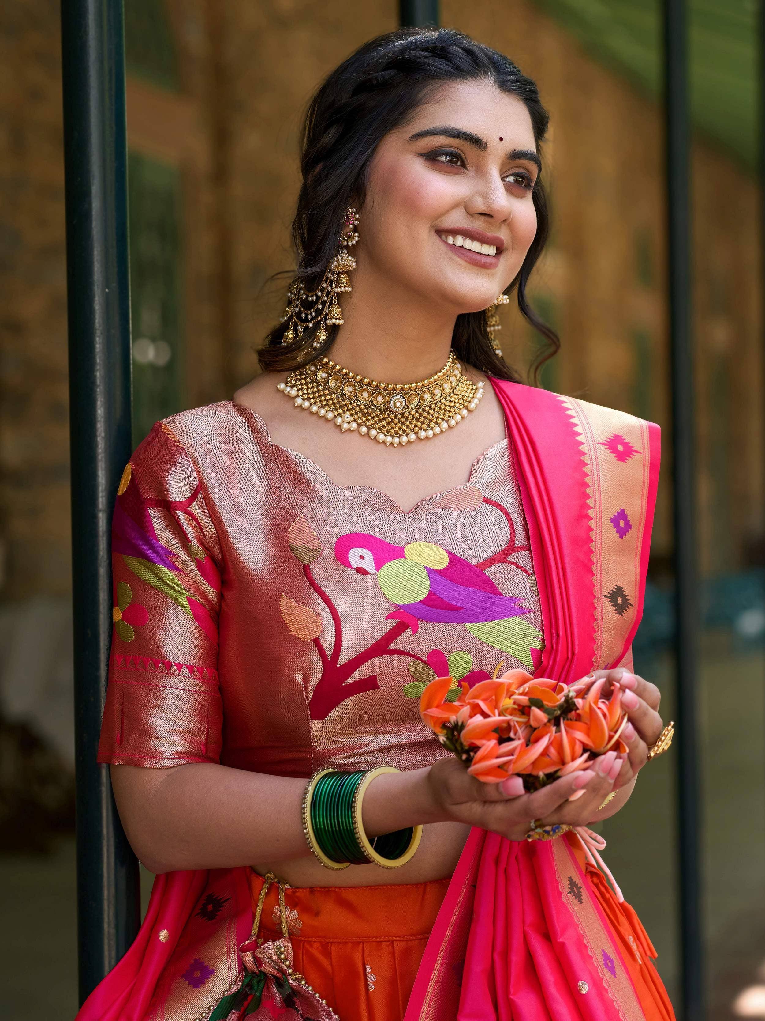 Woman in vibrant Indian ethnic wear with floral print blouse, pink dupatta, and gold jewelry holding orange flowers