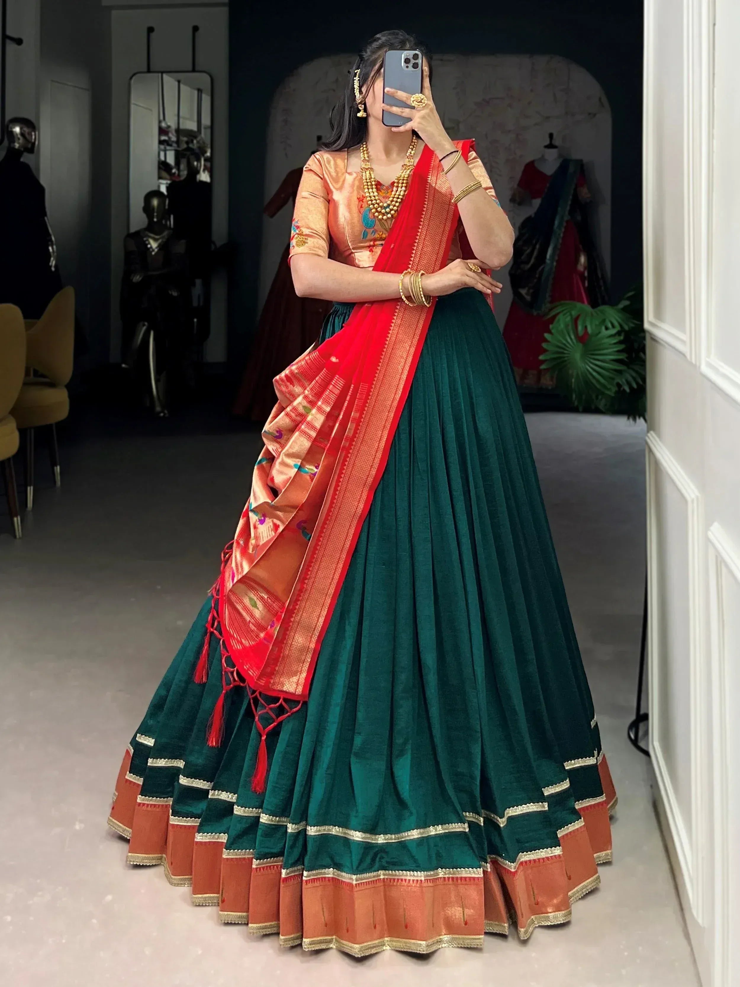 Woman in emerald green lehenga with red dupatta, gold jewelry, taking mirror selfie in boutique