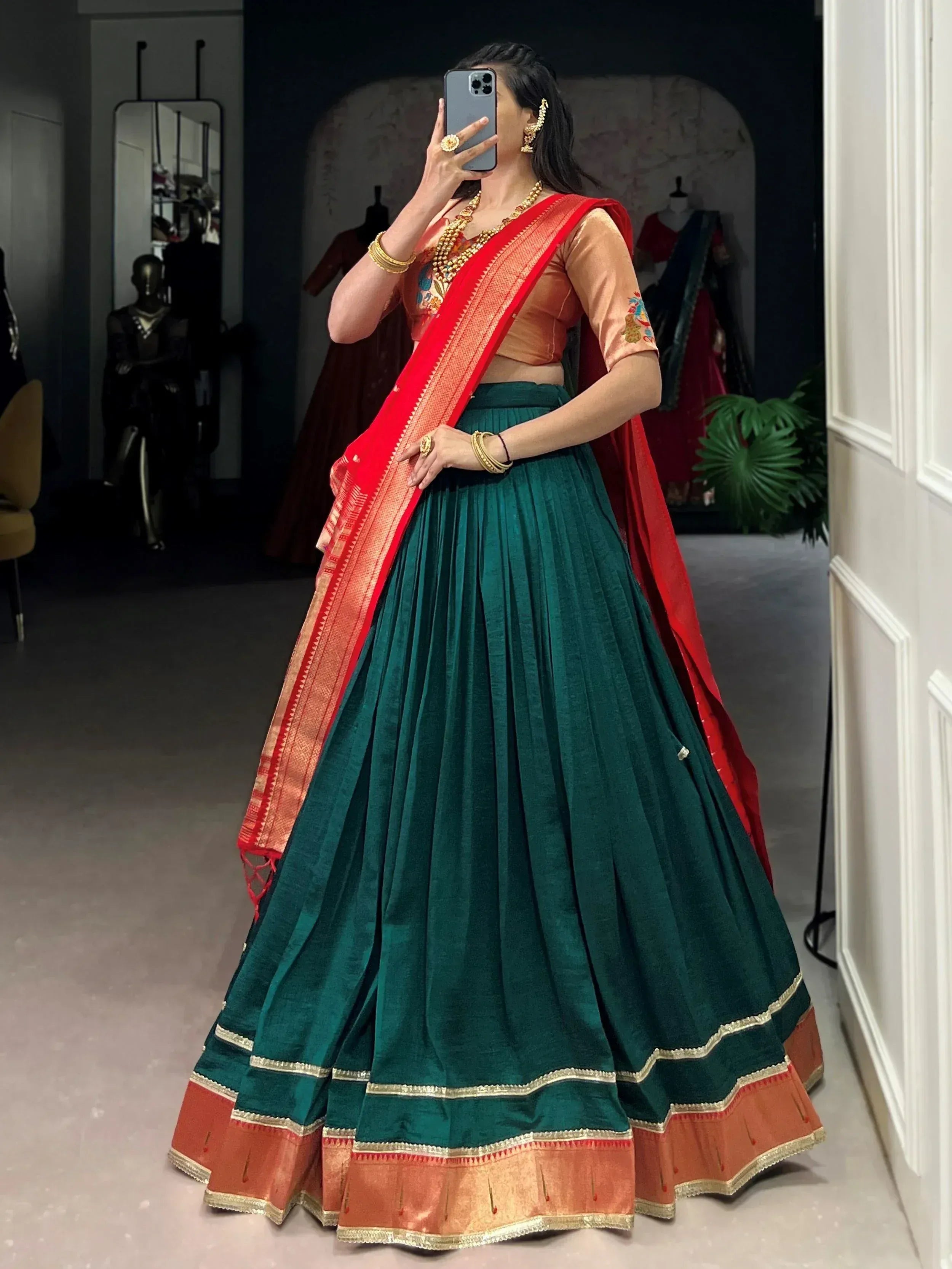 Woman in green velvet lehenga choli with red dupatta, gold jewelry, taking mirror selfie in boutique