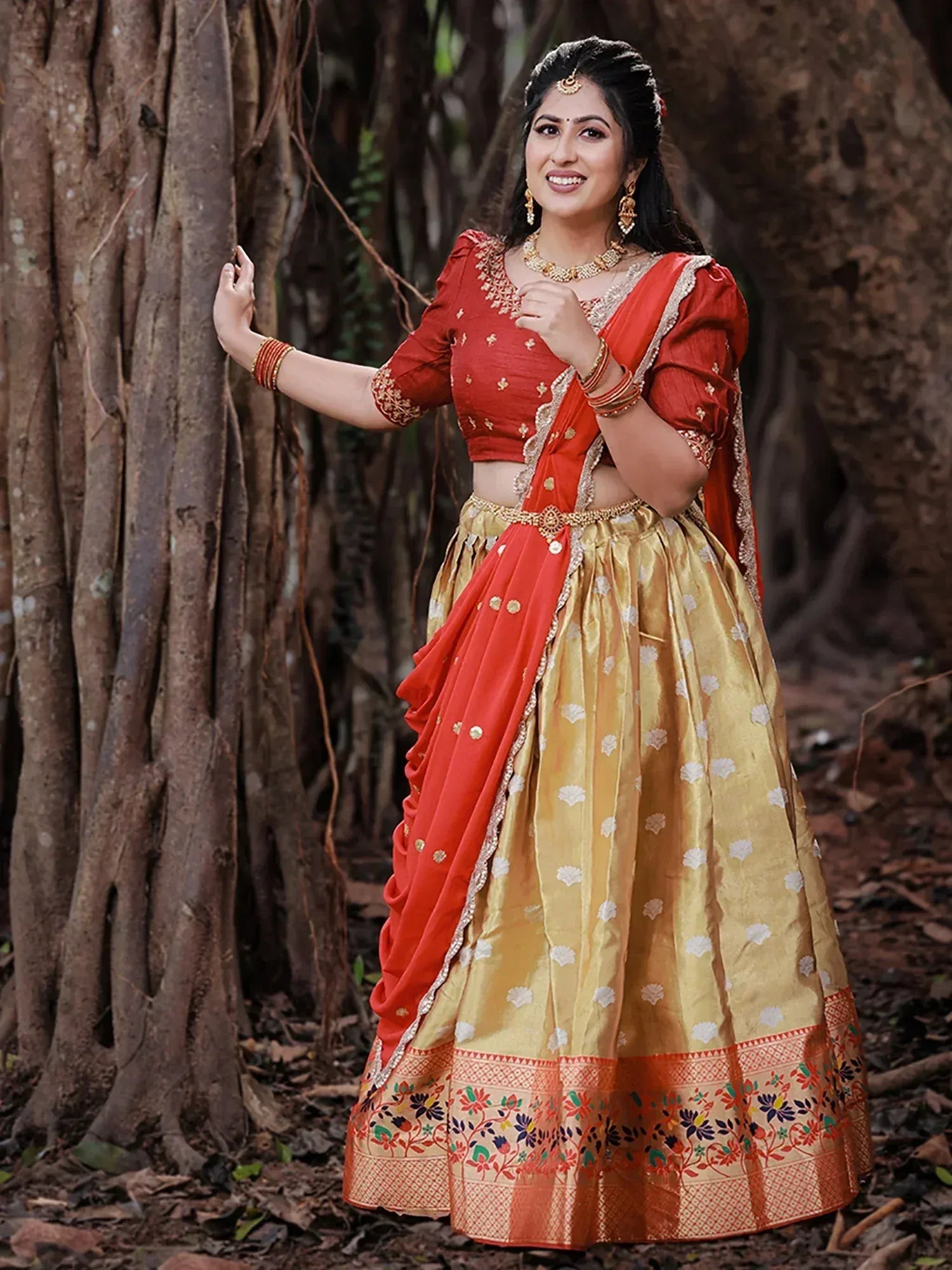 Woman in red and gold designer lehenga choli with floral motifs, standing by tree in forest setting