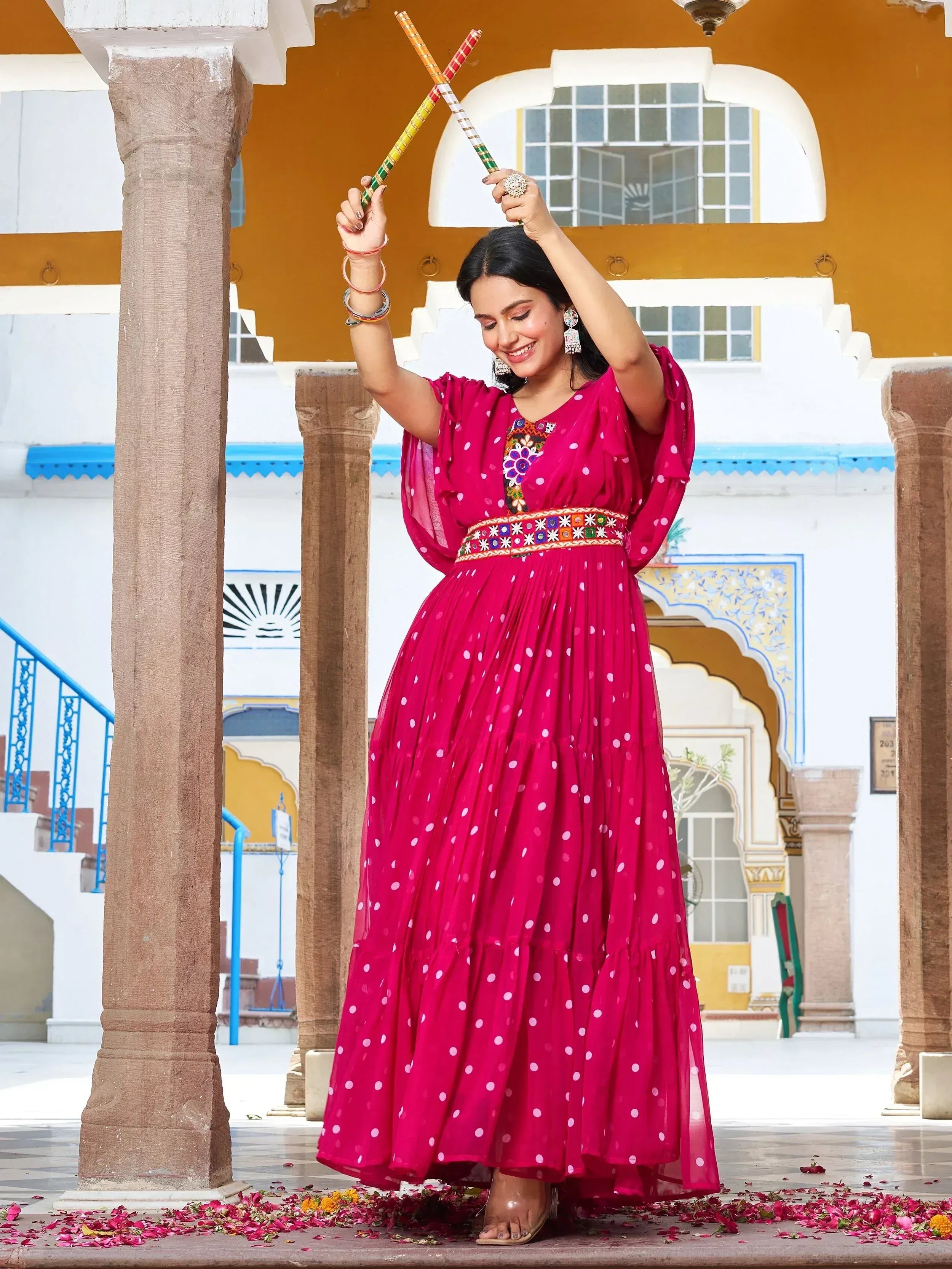 Woman in vibrant pink polka dot ethnic gown with mirror work, dancing with dandiya sticks in traditional courtyard