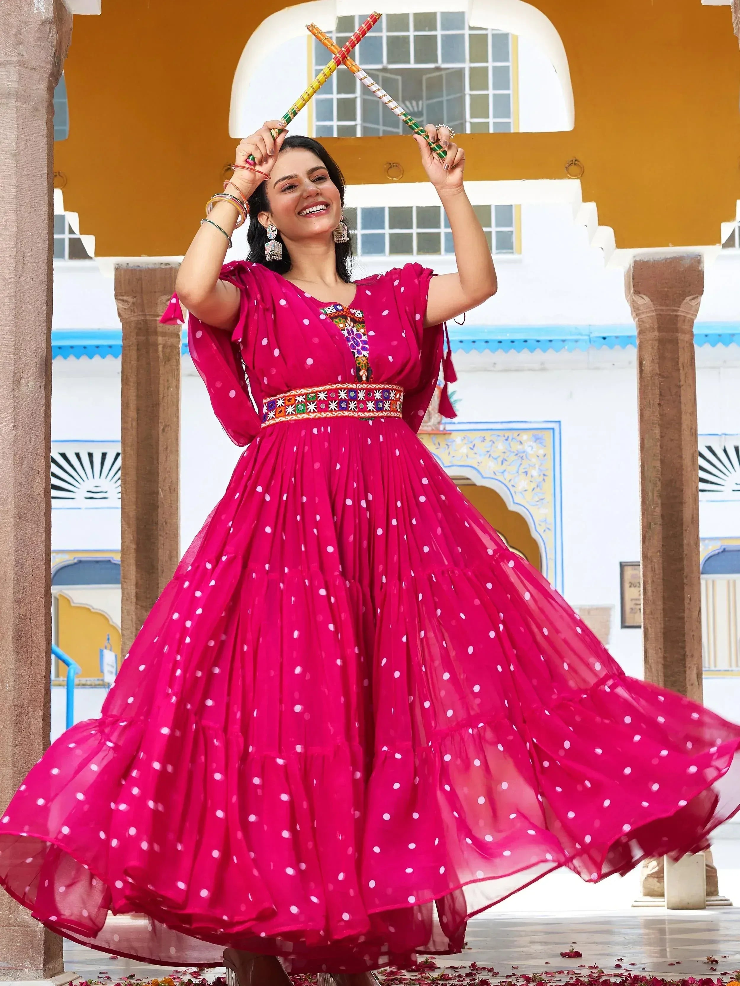 Woman in vibrant pink polka dot ethnic gown with mirror work, dancing with dandiya sticks
