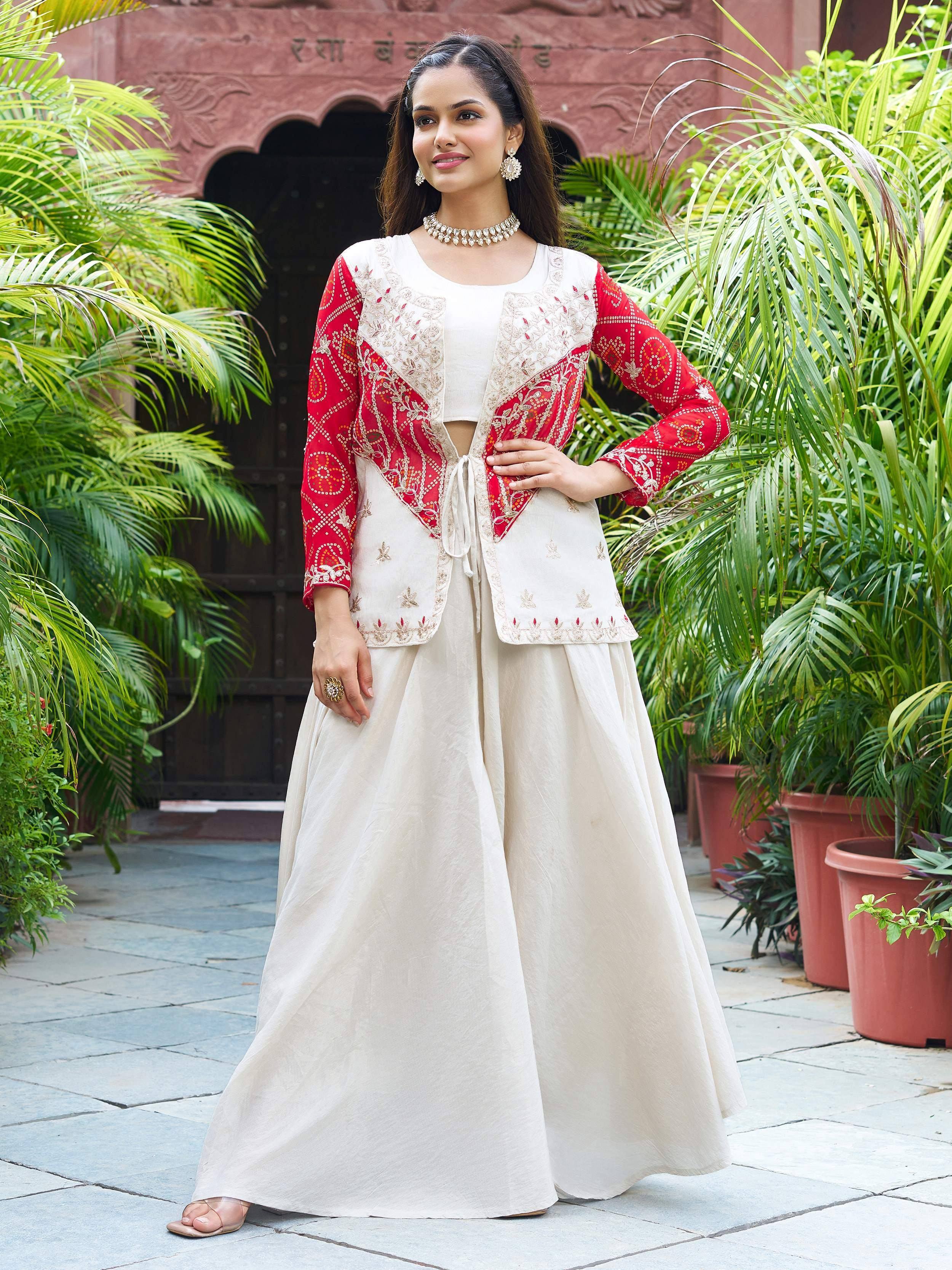 Woman in cream and red embroidered ethnic Indian outfit with floral patterns, standing outdoors near greenery.
