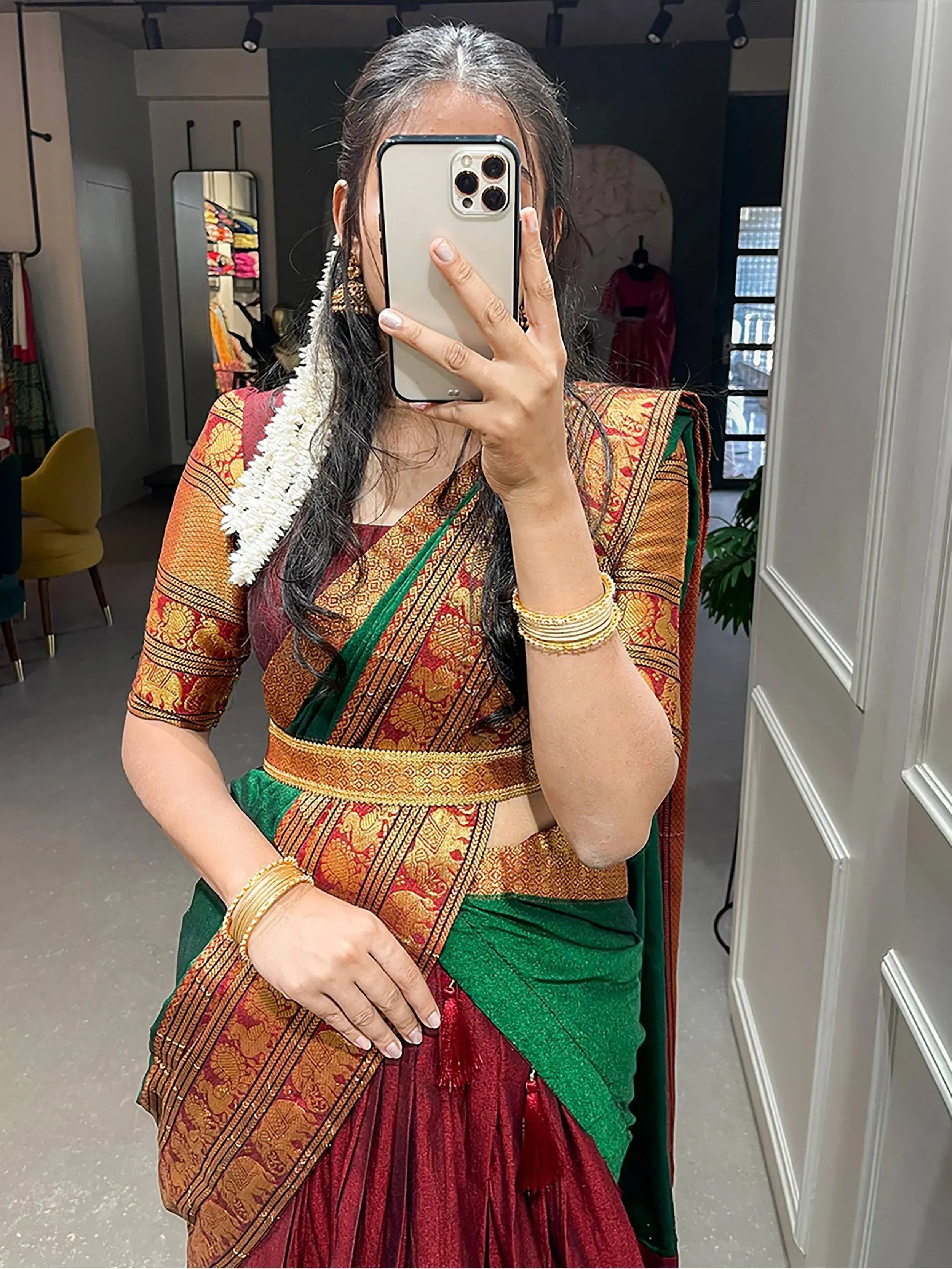 Woman in traditional Indian saree with gold embroidery, green border, and floral hair accessory taking mirror selfie