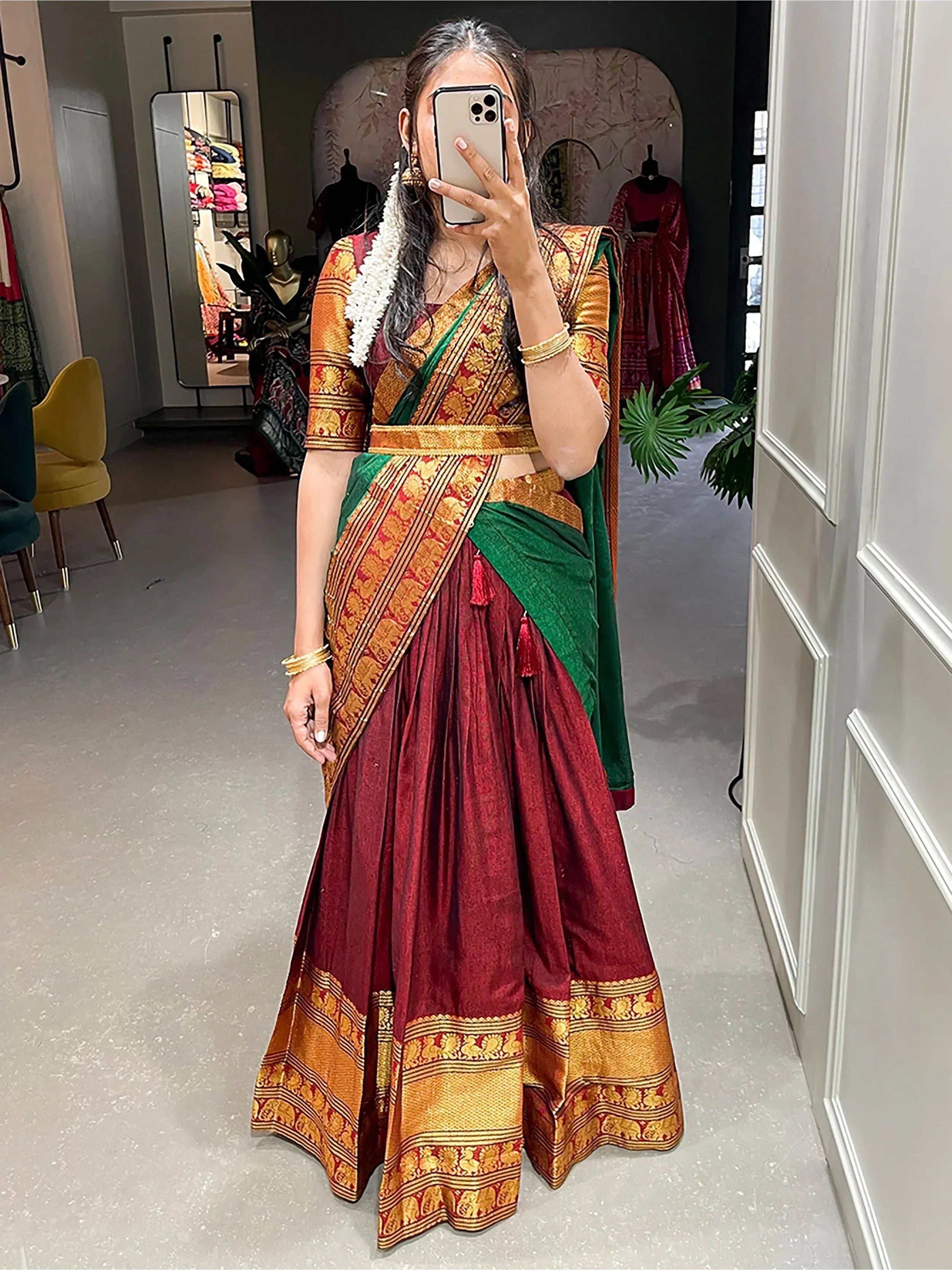 Woman in maroon and green designer lehenga choli with gold embroidery, taking mirror selfie in boutique