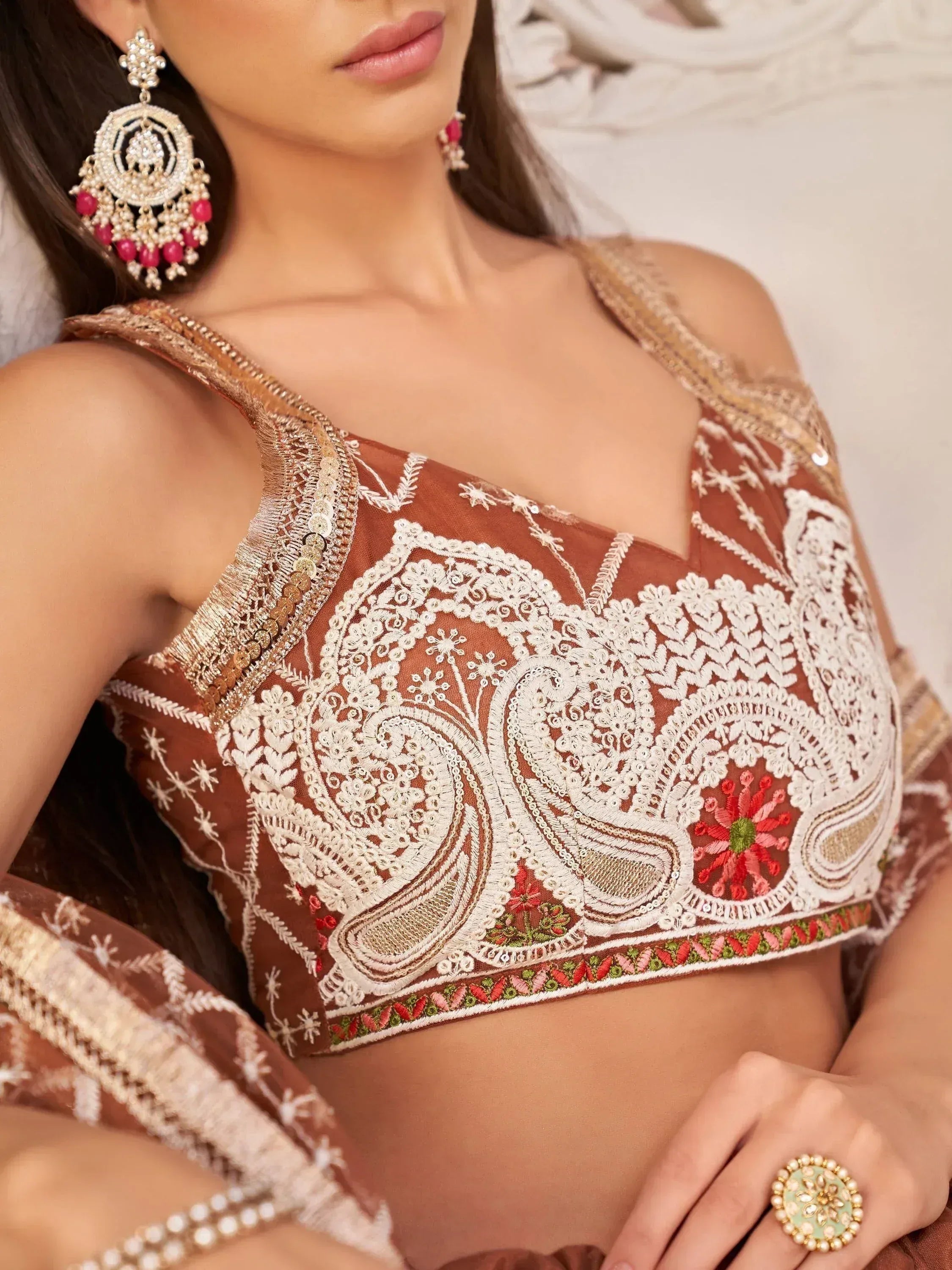Brown embroidered designer lehenga choli top with white paisley and floral motifs, paired with statement earrings.