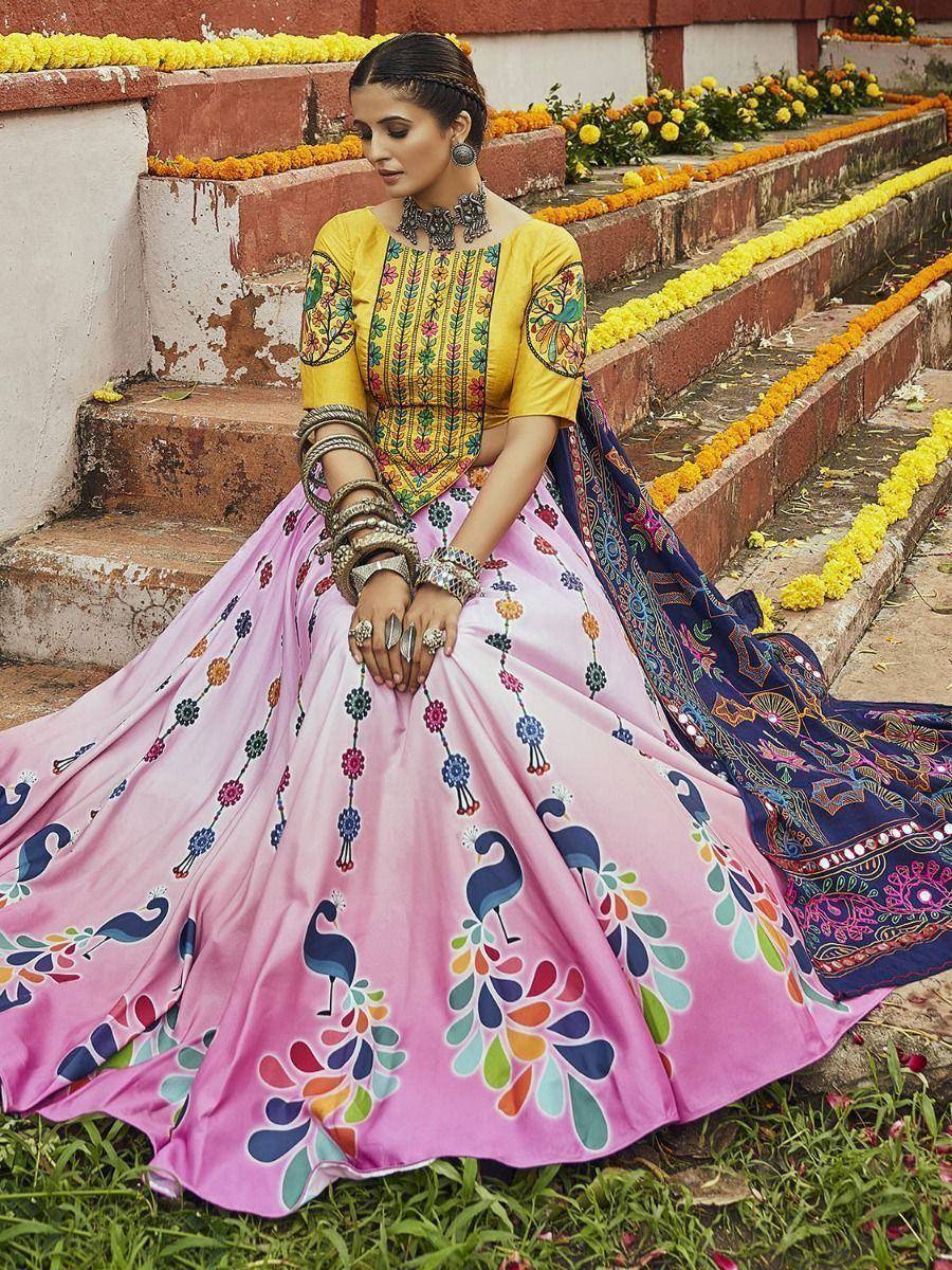Woman in vibrant yellow and pink embroidered lehenga choli with peacock print, styled with traditional jewelry and dupatta, seated on floral steps.