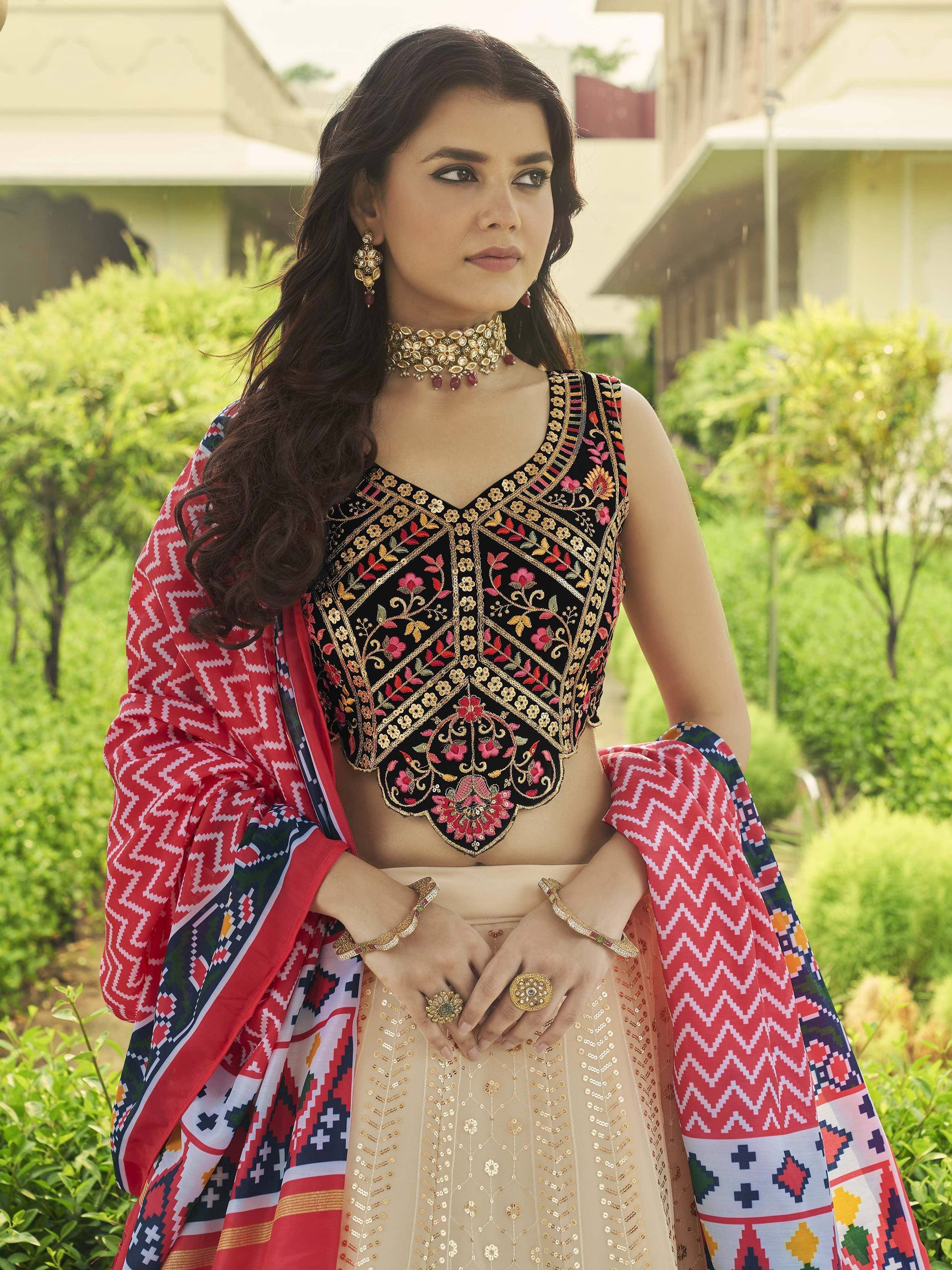 Woman in embroidered black crop top with floral motifs, beige sequined skirt, and red patterned dupatta, styled with gold jewelry for Indian ethnic wear.