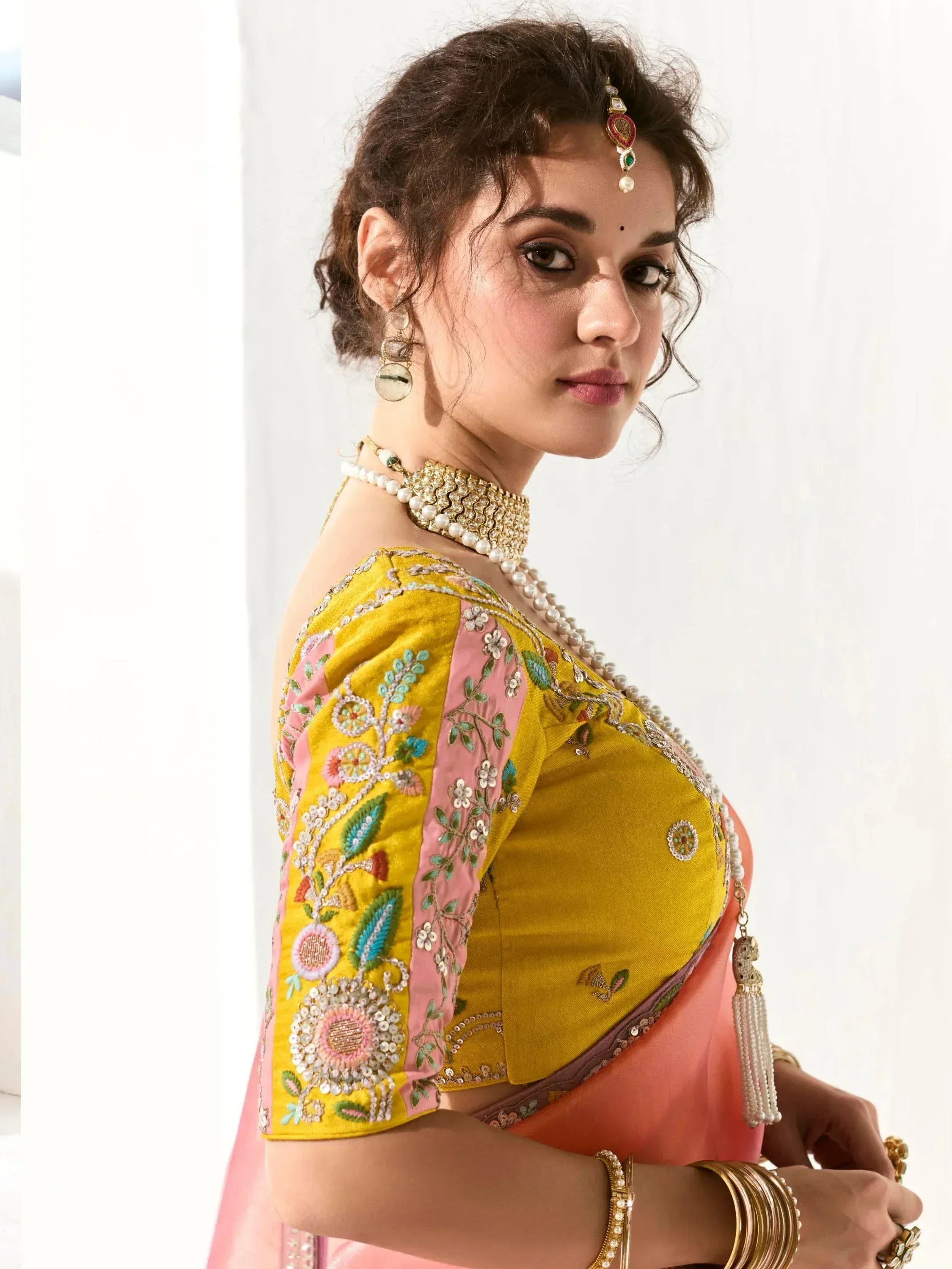 Yellow and pink embroidered saree blouse with floral detailing, paired with traditional jewelry for Indian ethnic wear.