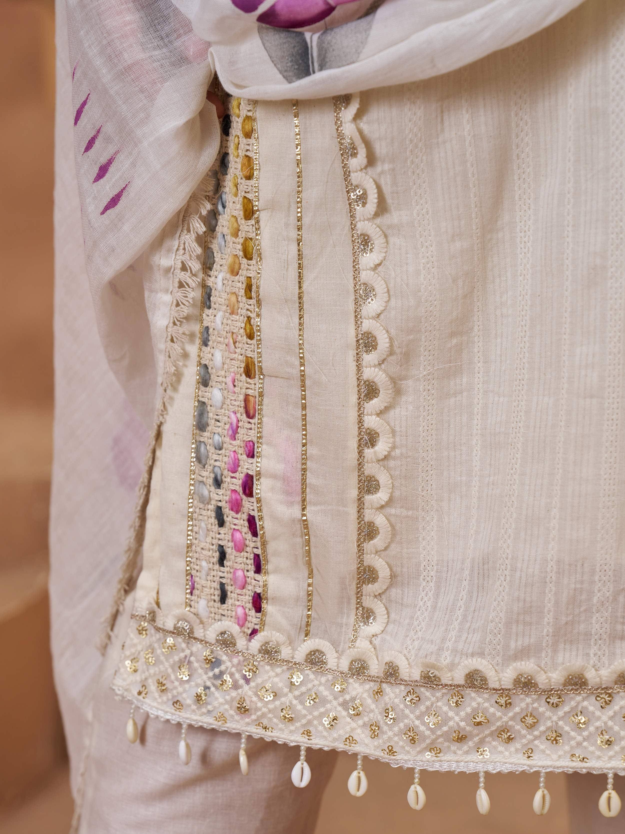 Cream ethnic kurti with colorful threadwork, gold trim, and cowrie shell tassels, paired with floral dupatta.