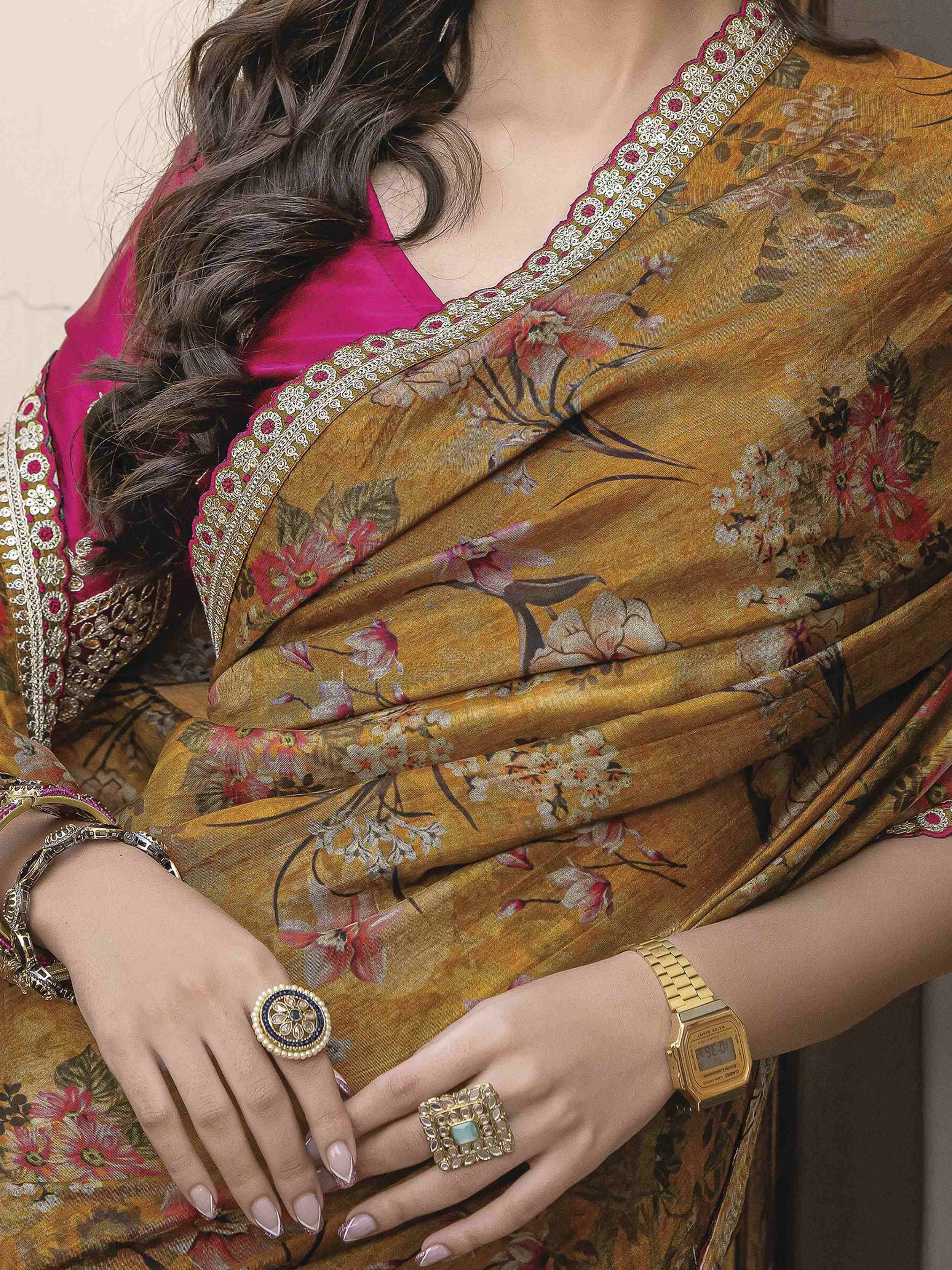Woman in floral mustard saree with pink blouse, ethnic jewelry, and gold watch, showcasing Indian ethnic wear.
