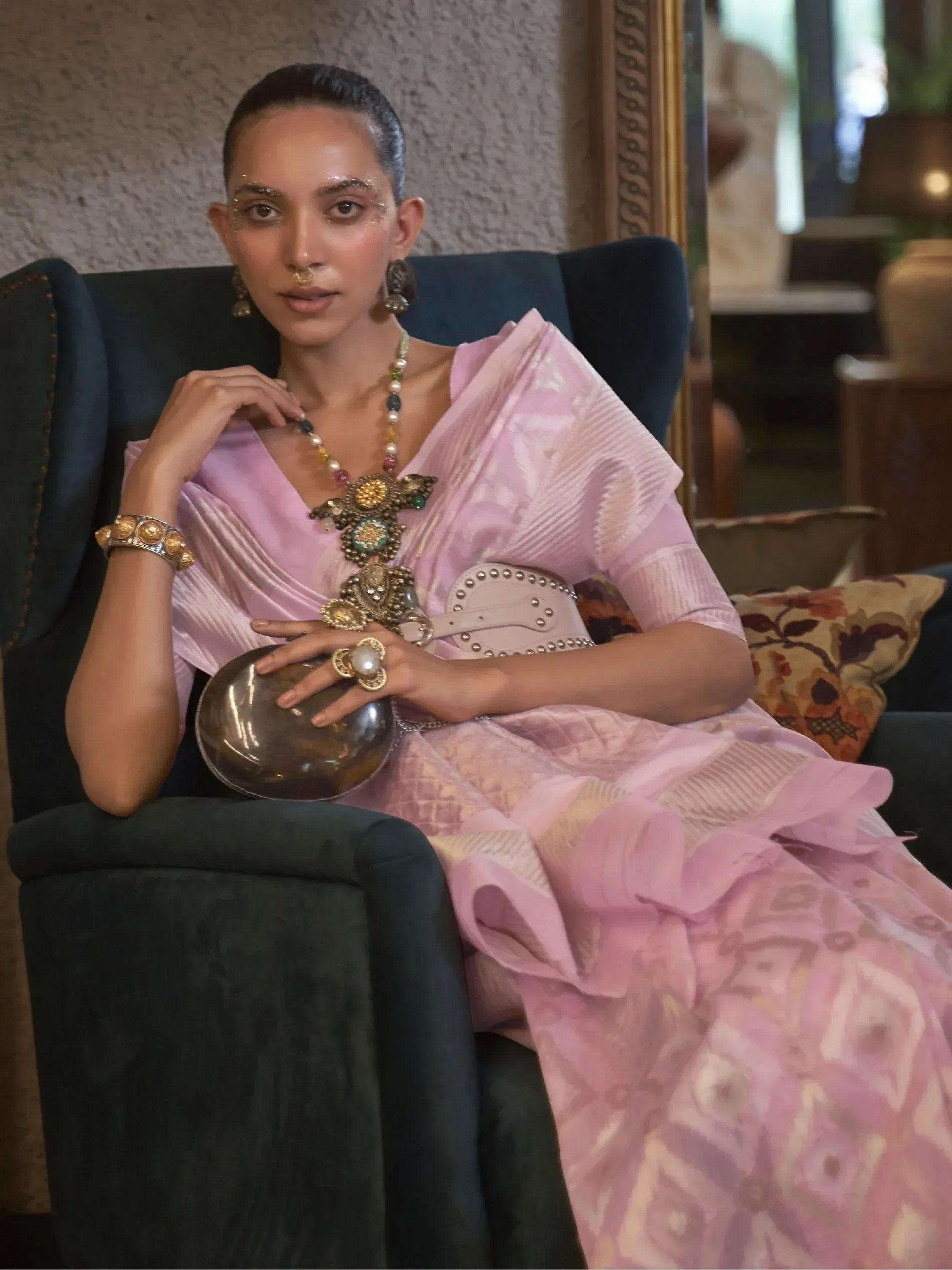 Woman in pink ethnic gown with statement jewelry, seated on velvet chair