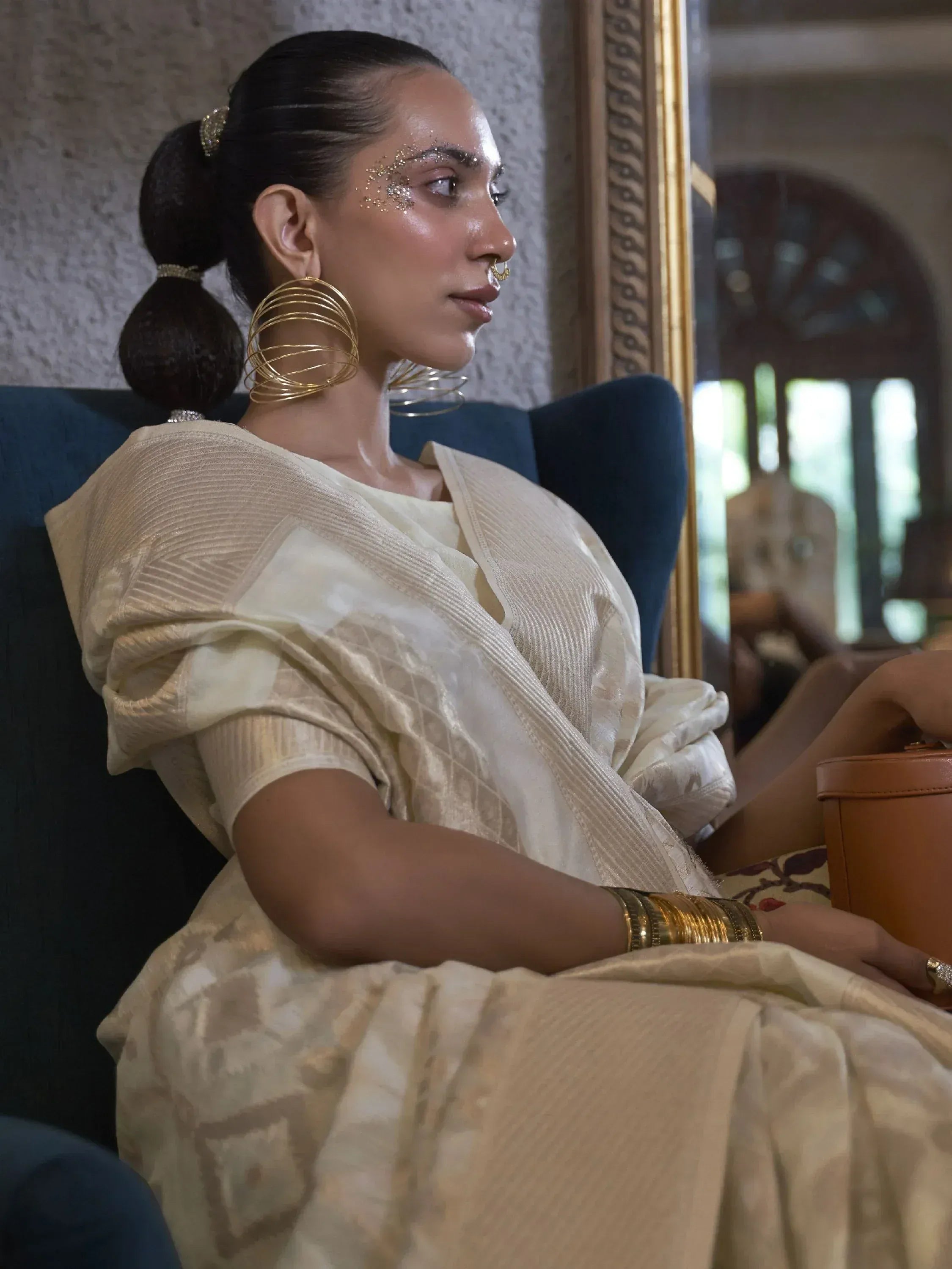 Elegant woman in cream ethnic saree with gold jewelry, sitting on blue chair, festive makeup