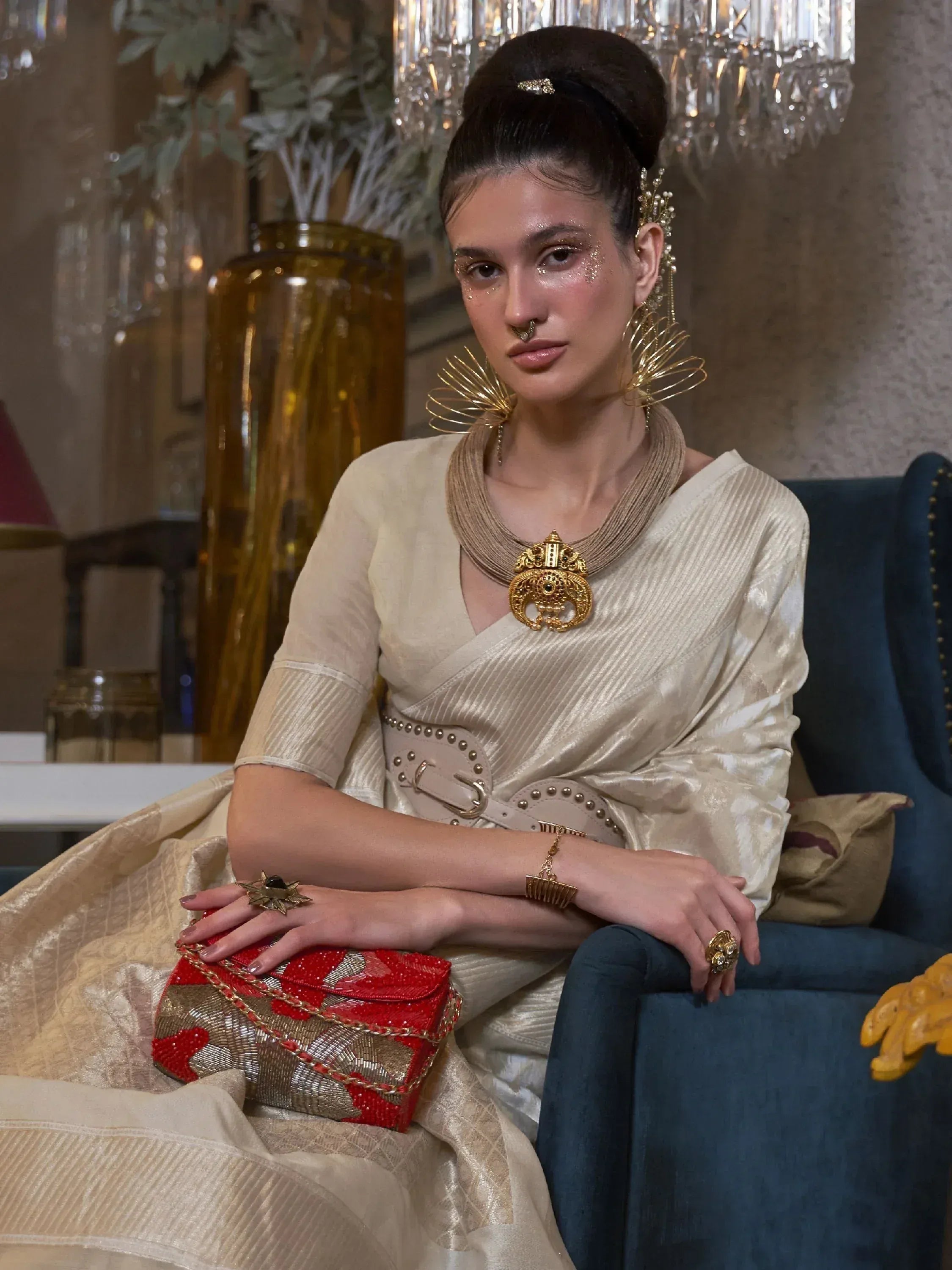 Elegant woman in cream ethnic gown with gold jewelry and red clutch, styled for festive occasions.