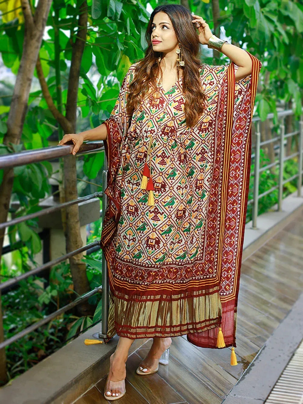 Woman in printed ethnic kaftan gown with tassels, standing on wooden deck amidst greenery