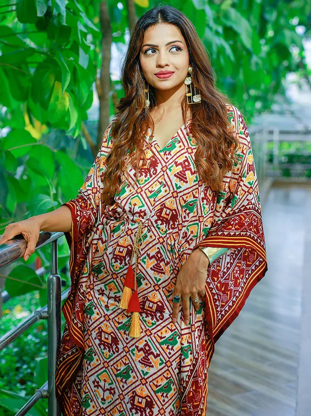 Woman in colorful printed ethnic gown with tassels, standing by railing amidst greenery