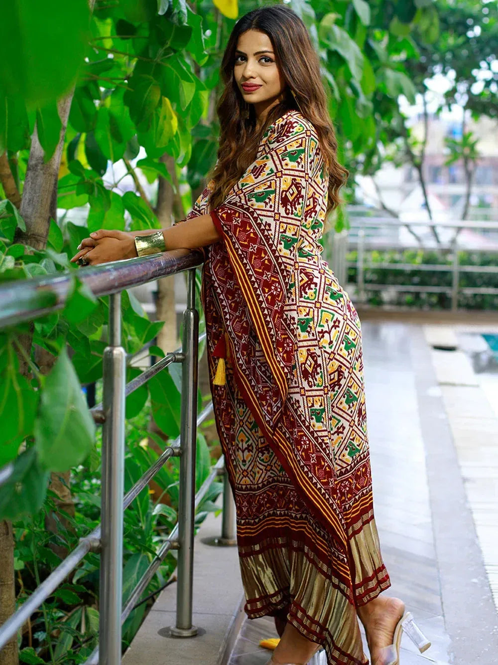 Woman in colorful printed ethnic gown with geometric patterns, standing by railing amidst greenery