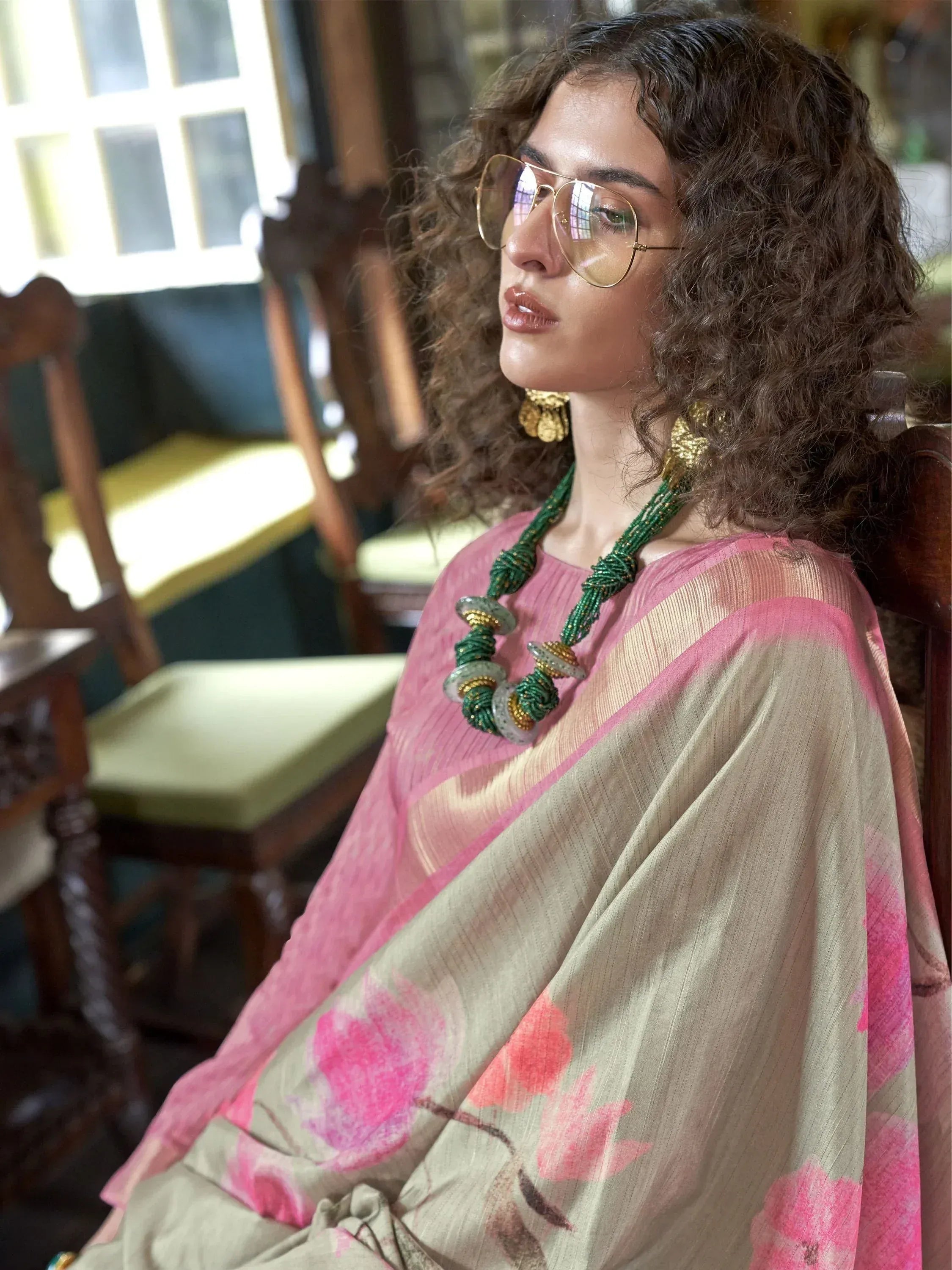 Woman in pink floral saree with green beaded necklace, gold earrings, and sunglasses, seated indoors.