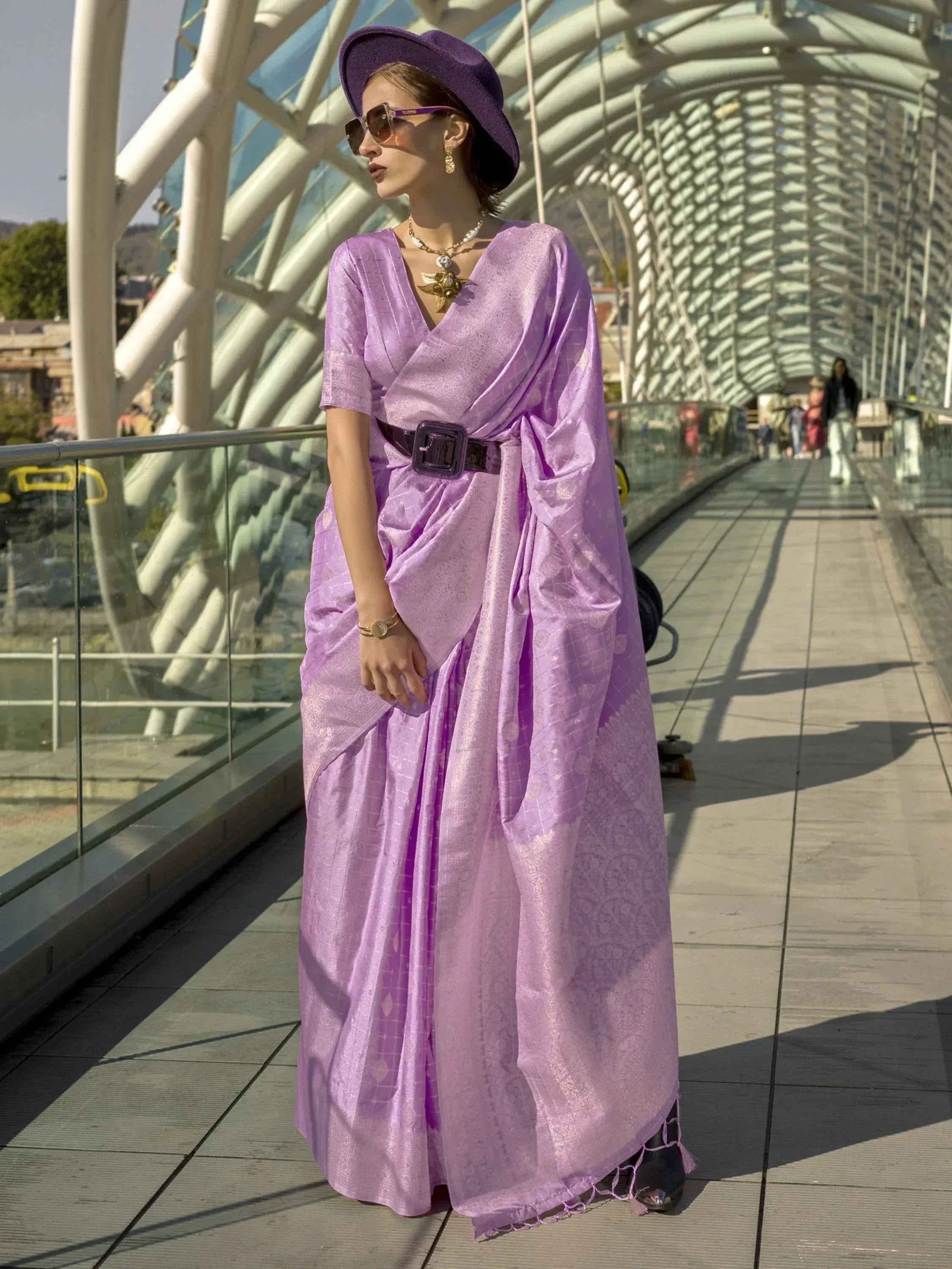 Woman in purple ethnic saree gown with hat, modern bridge backdrop, designer Indian wear