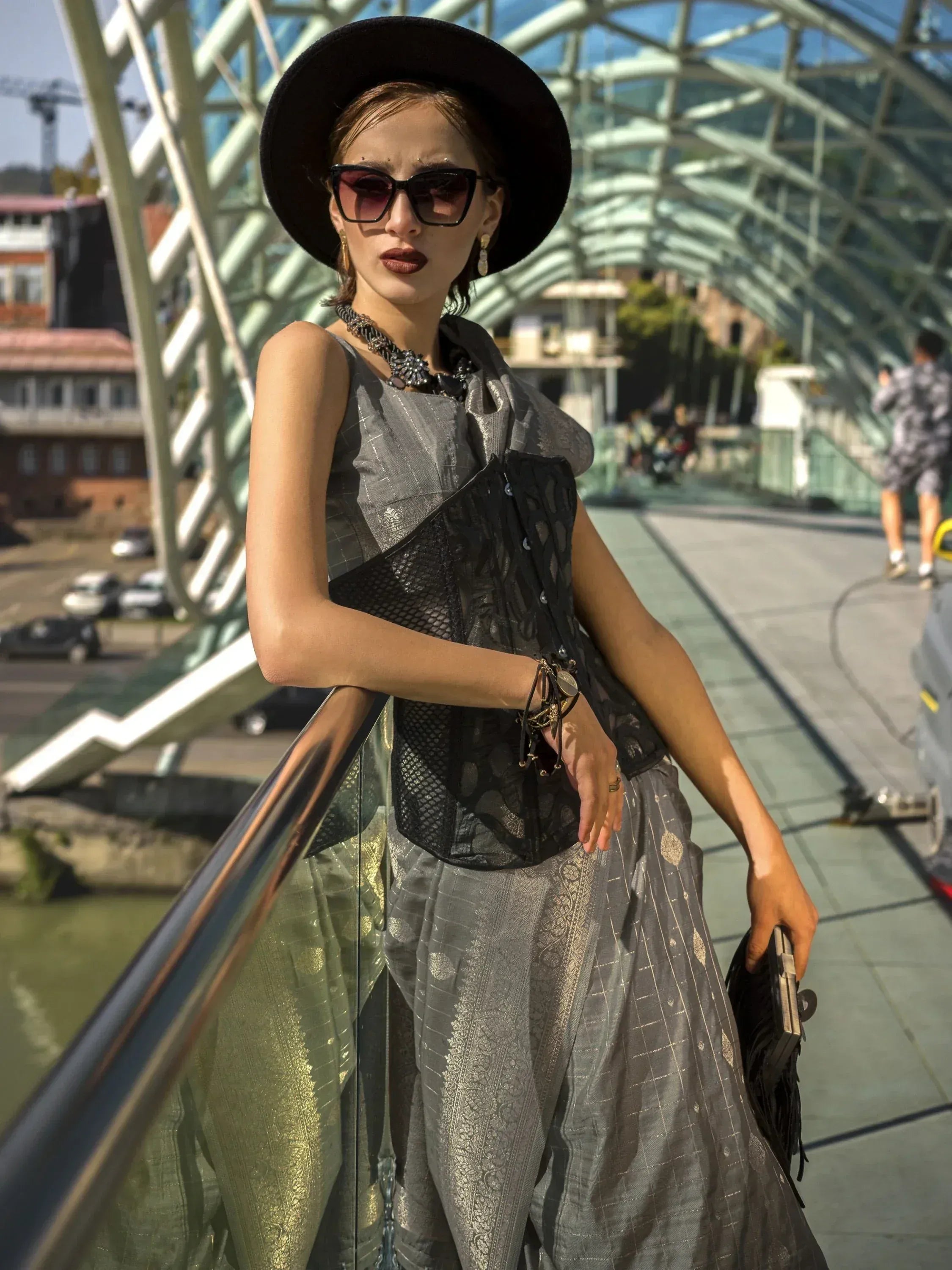 Stylish woman in silver ethnic gown with black lace top, hat, and sunglasses on modern bridge
