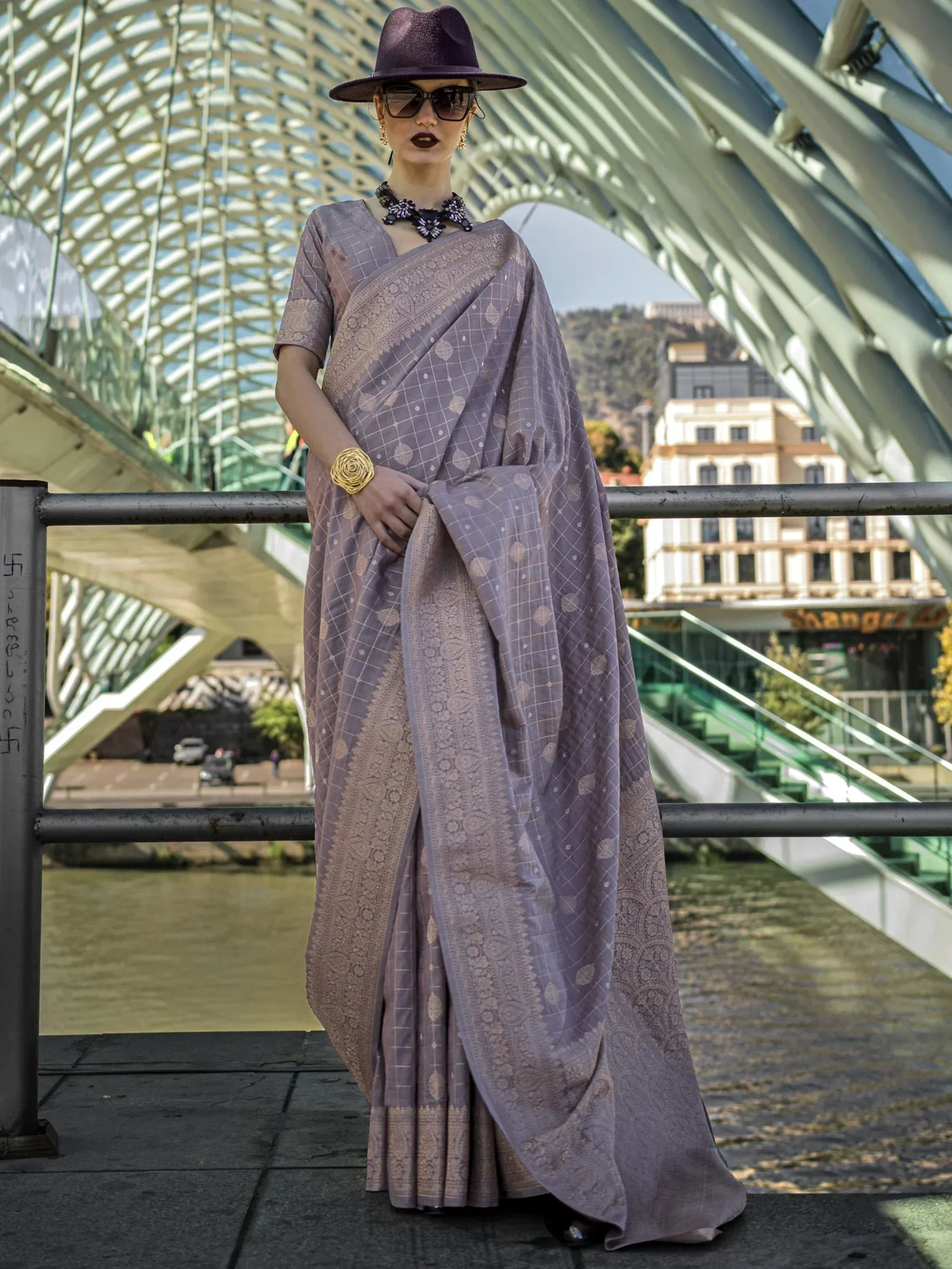 Stylish purple saree with gold embroidery, worn with hat and sunglasses on a modern bridge.