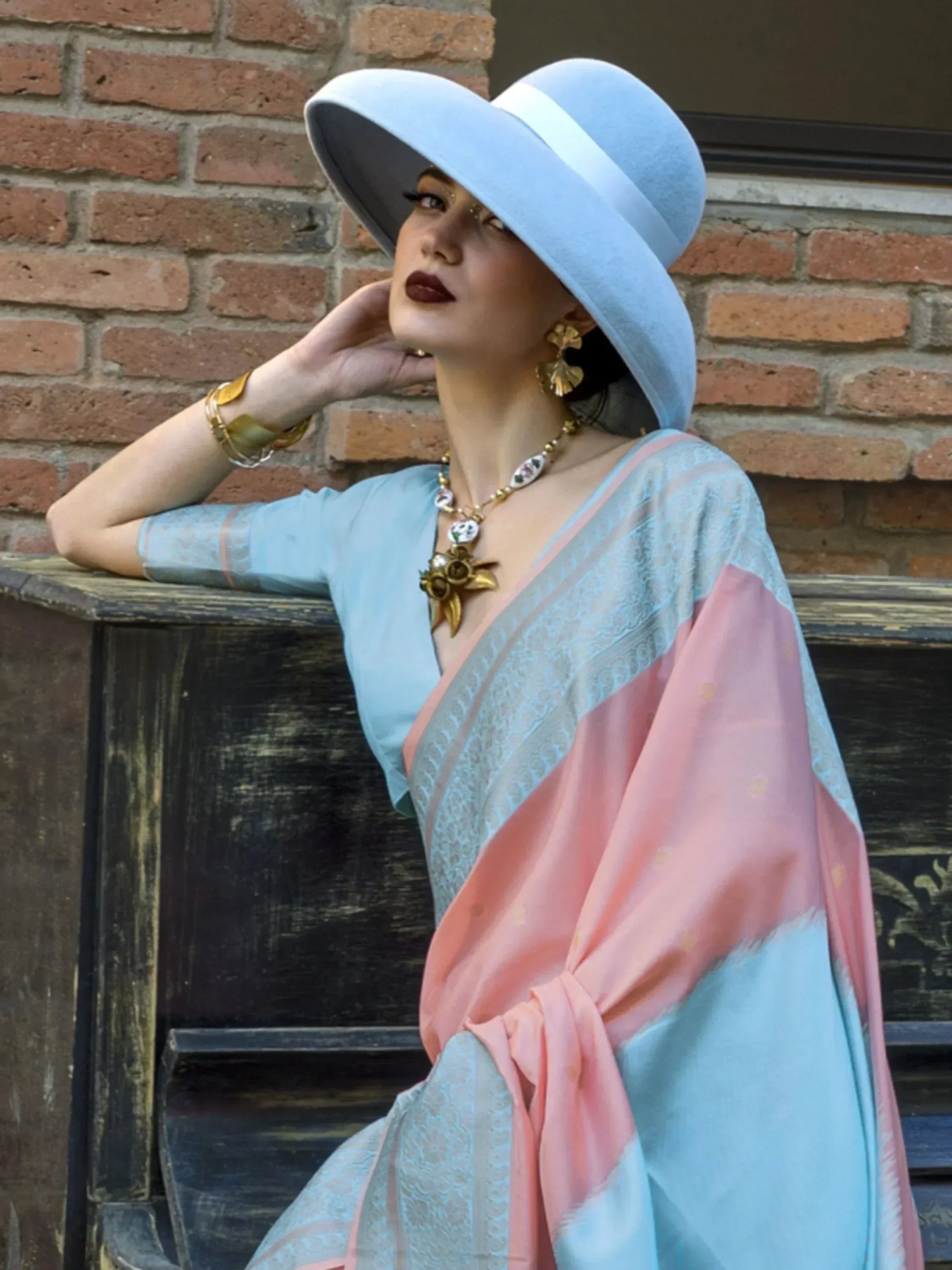 Woman in pastel blue and pink ethnic gown with wide-brim hat, gold jewelry, against brick wall