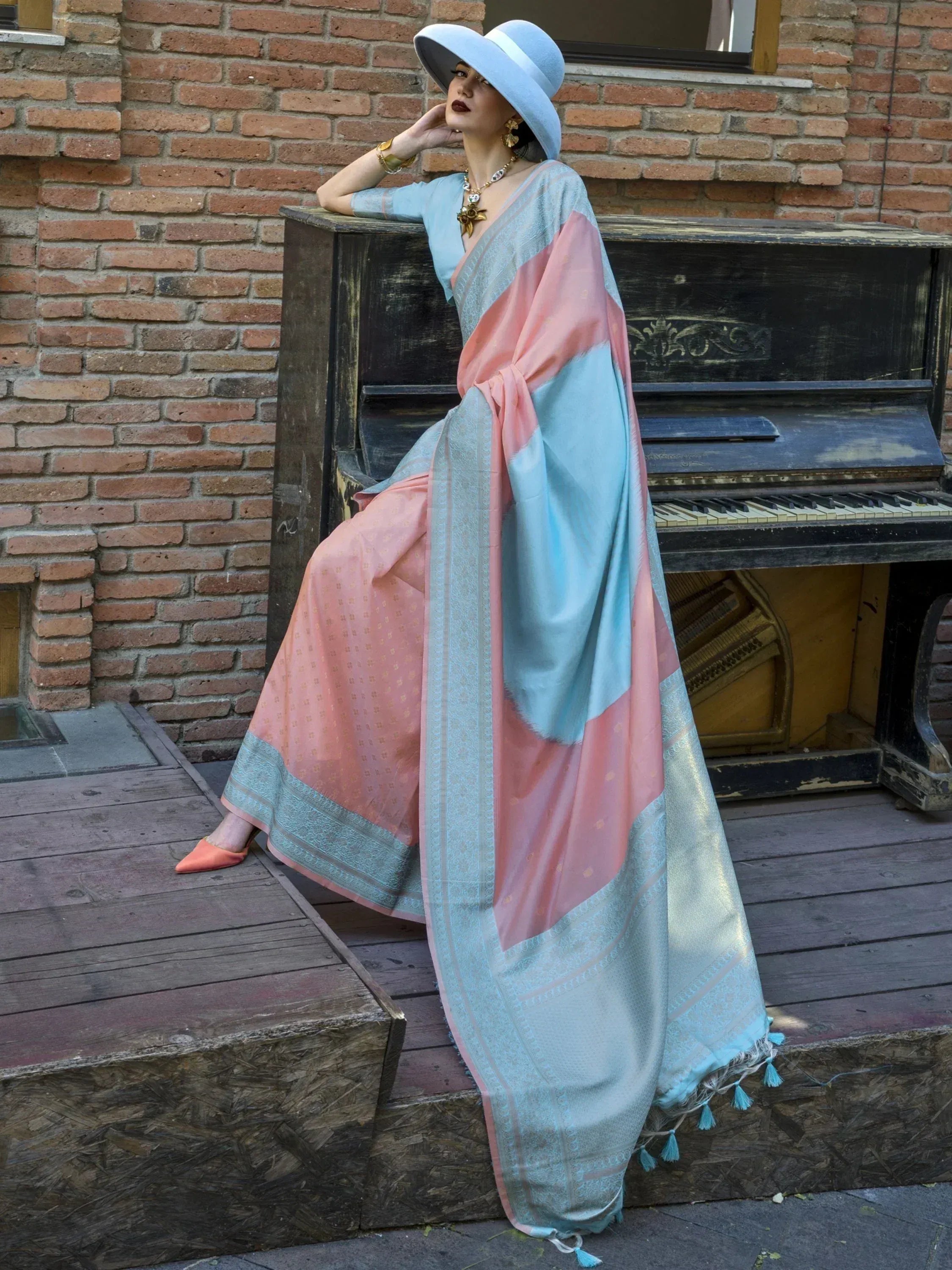 Woman in pastel pink and blue designer saree with gold motifs, styled with wide-brim hat, posing by vintage piano