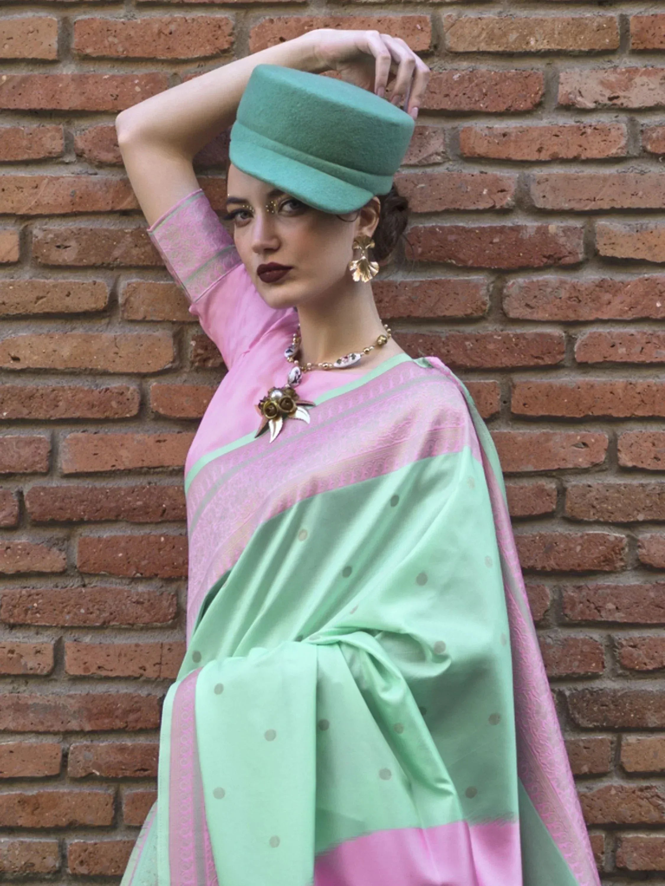 Woman in pink and mint green saree with matching hat, gold jewelry, against brick wall