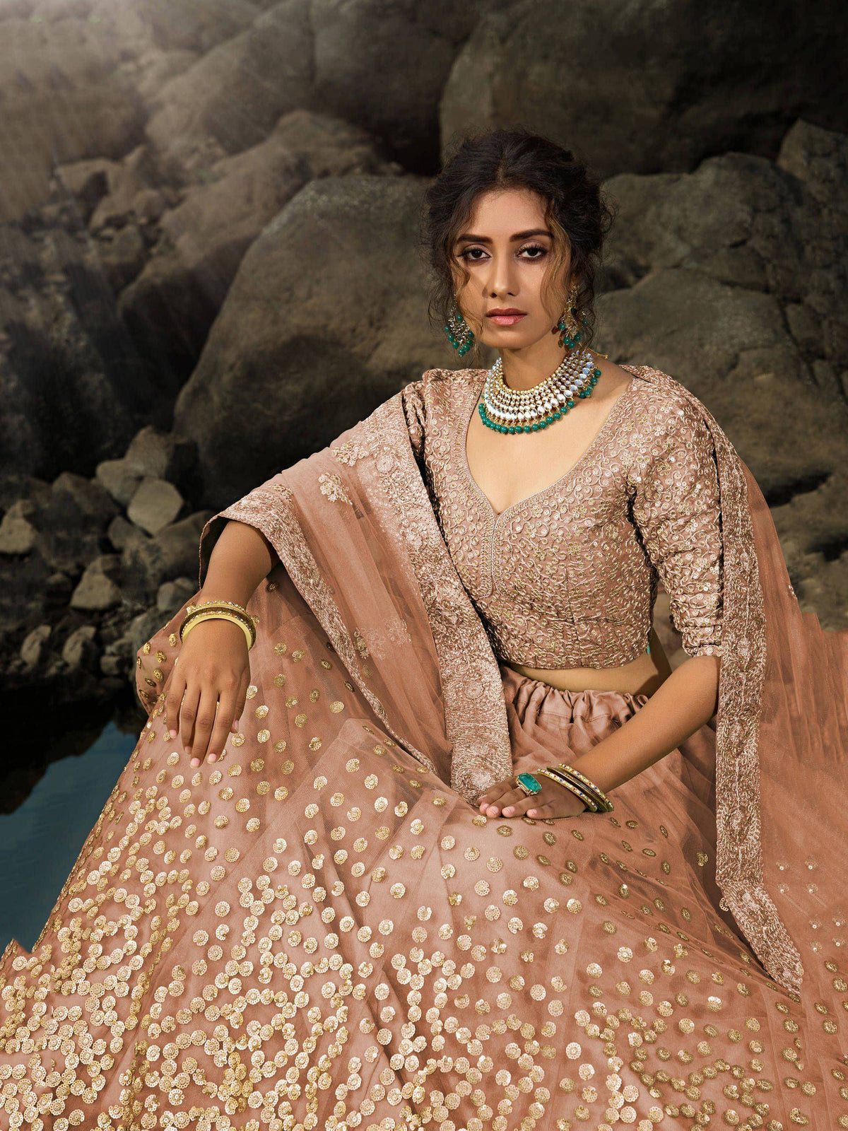 Woman in elegant blush pink embroidered lehenga choli with gold motifs, styled with turquoise jewelry against rocky backdrop