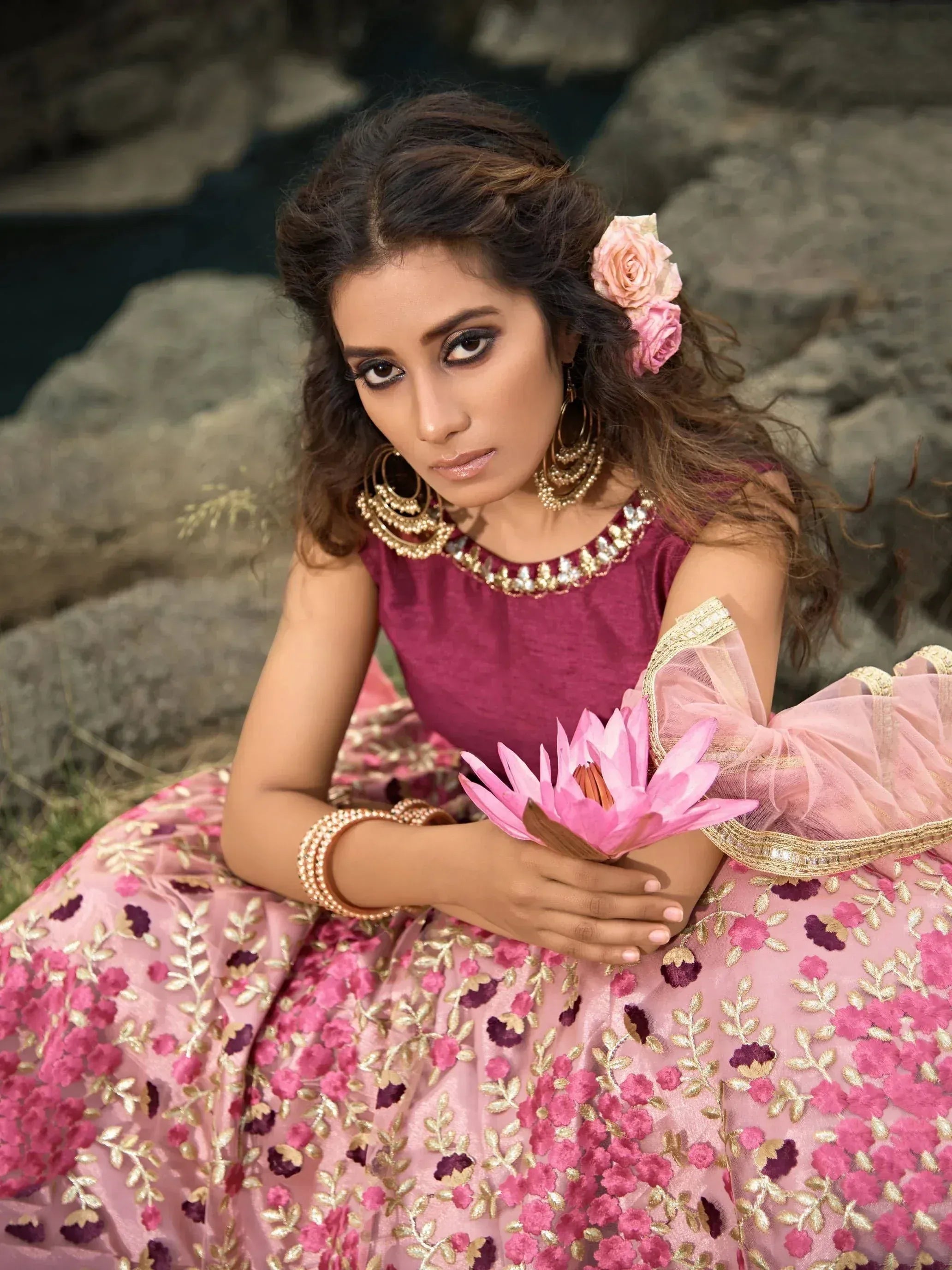 Woman in pink floral embroidered lehenga choli with gold jewelry, holding lotus flower, styled for Indian ethnic wear.