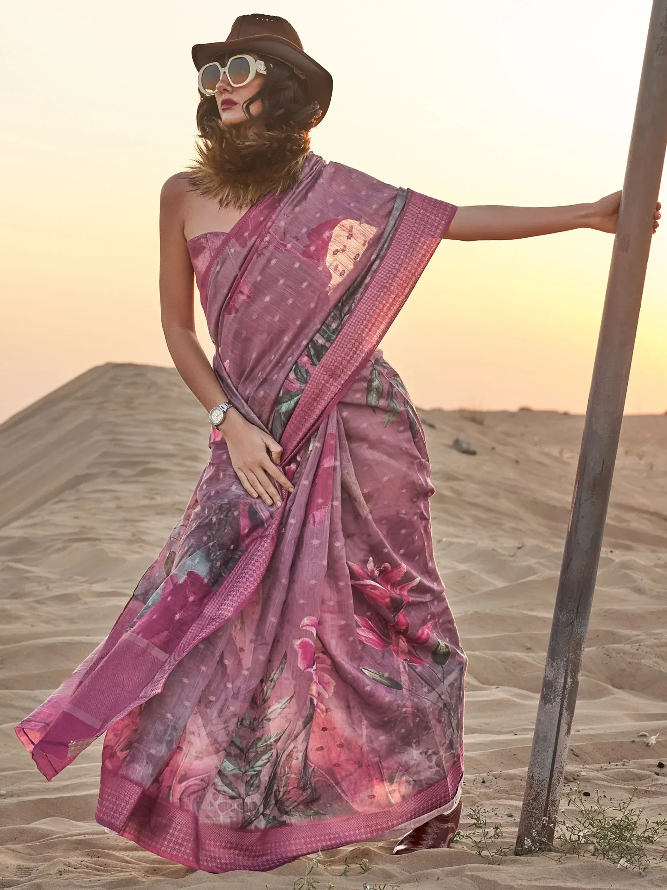 Woman in floral pink saree with hat and sunglasses, desert sunset backdrop, ethnic wear style
