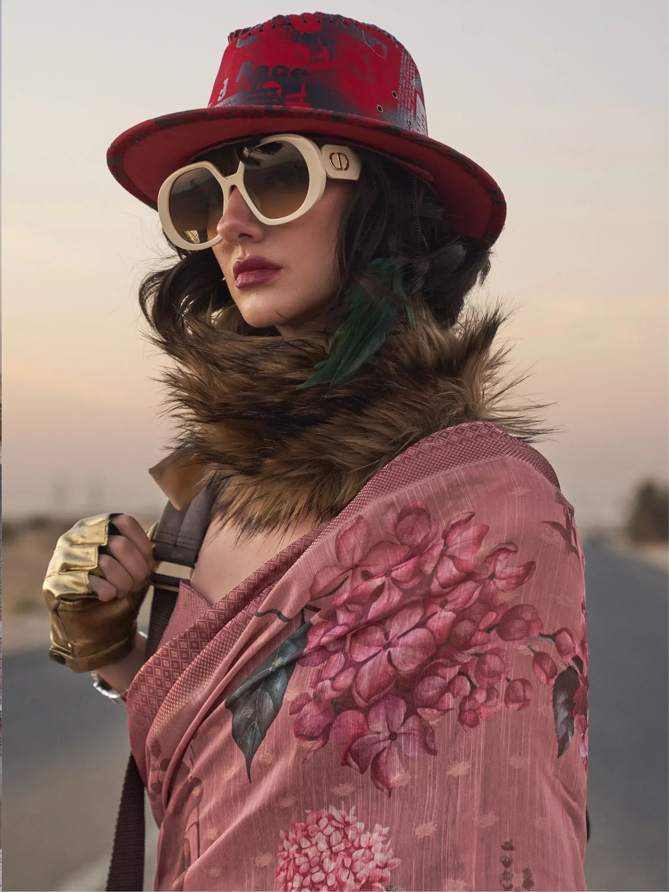 Woman in floral pink ethnic gown with fur collar, red hat, and sunglasses, styled for modern Indian fashion.