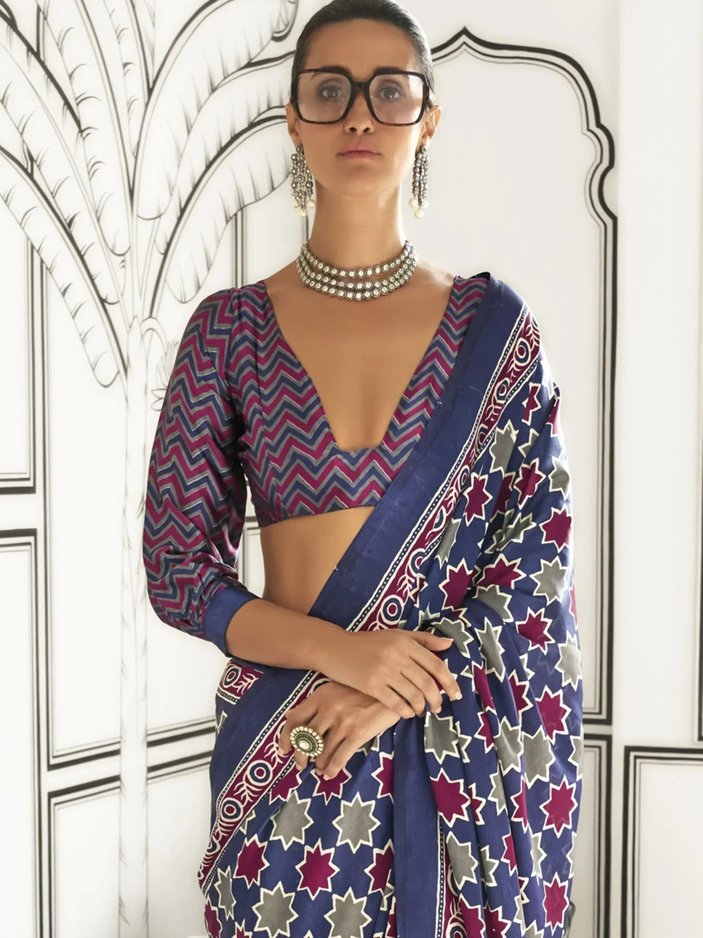 Woman in blue and pink printed saree with zigzag blouse, ethnic jewelry, and glasses