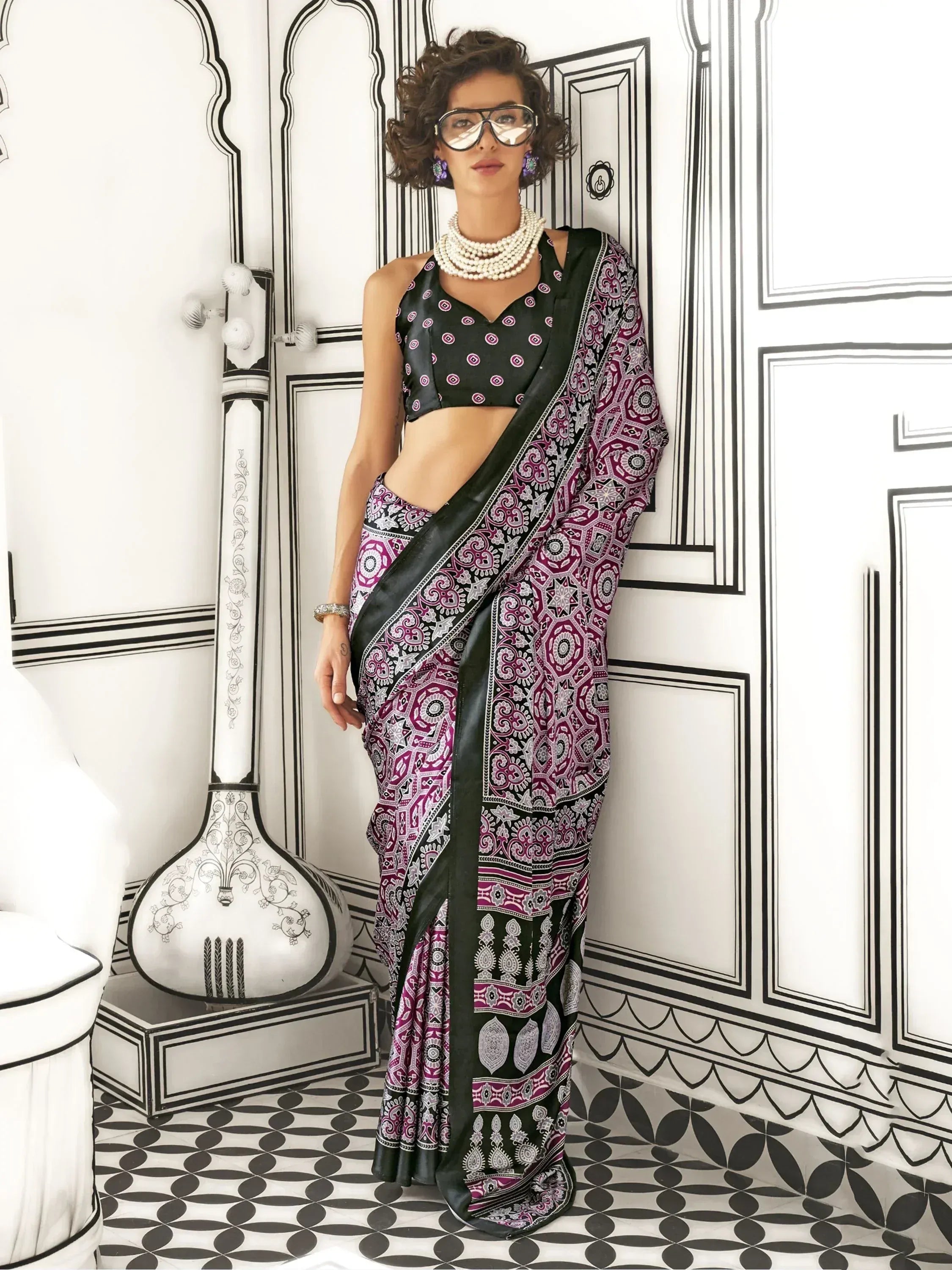 Stylish woman in purple and black printed saree with designer blouse, pearl necklace, and statement glasses.