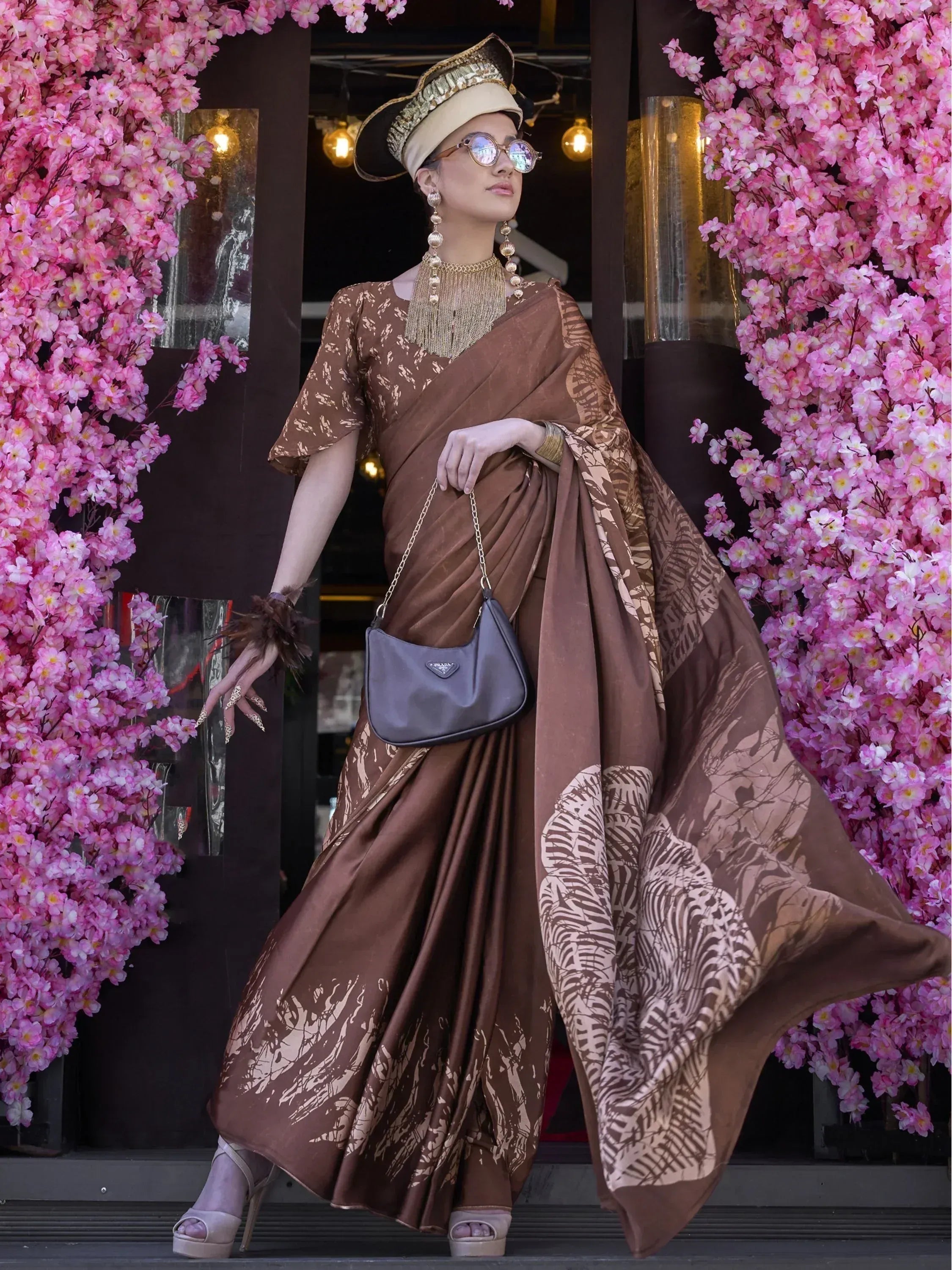 Stylish brown printed saree with gold accents, paired with statement jewelry and hat, perfect for ethnic occasions.