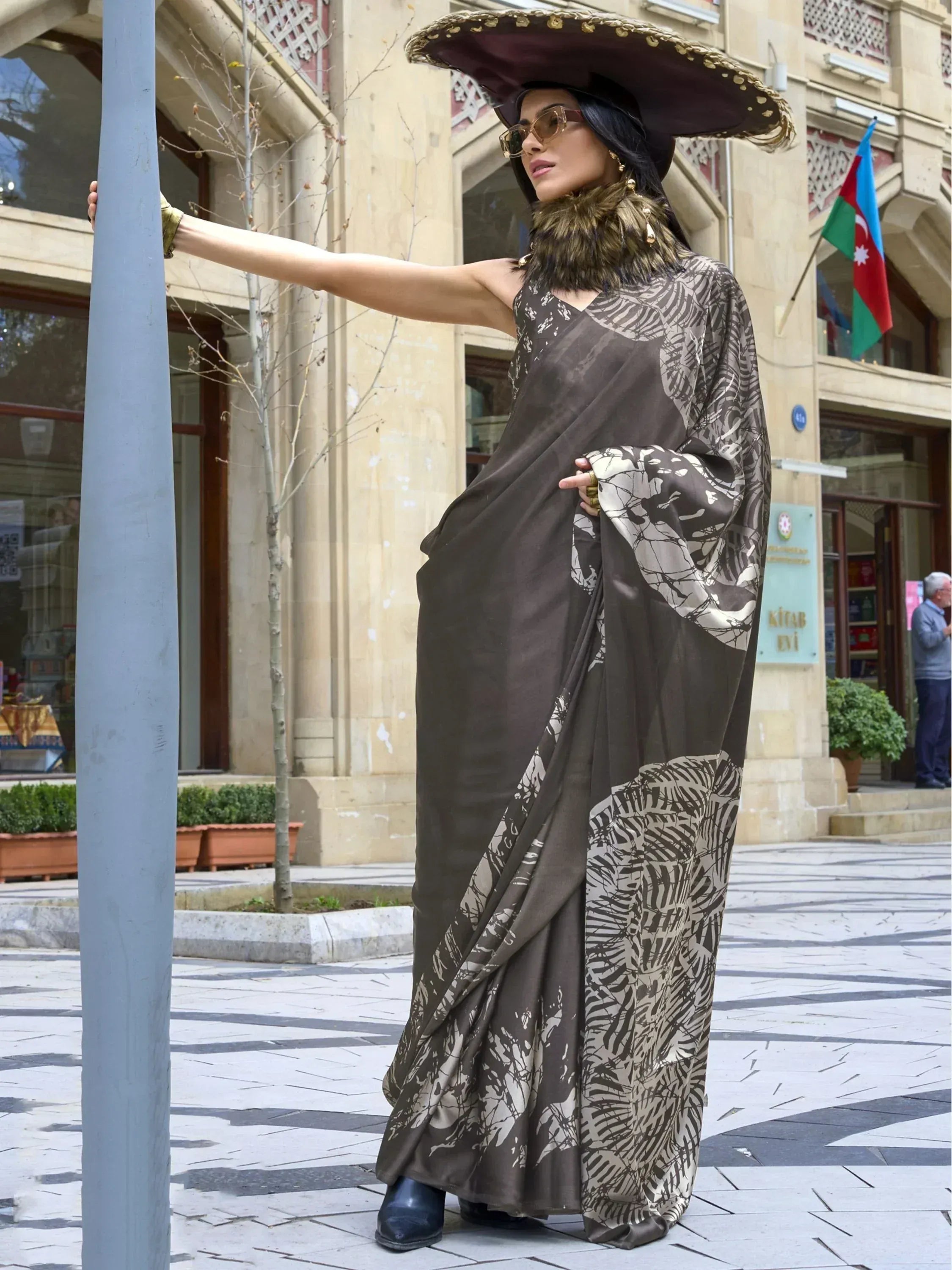 Woman in brown printed saree with wide-brim hat, fur collar, and sunglasses, styled for ethnic fashion.