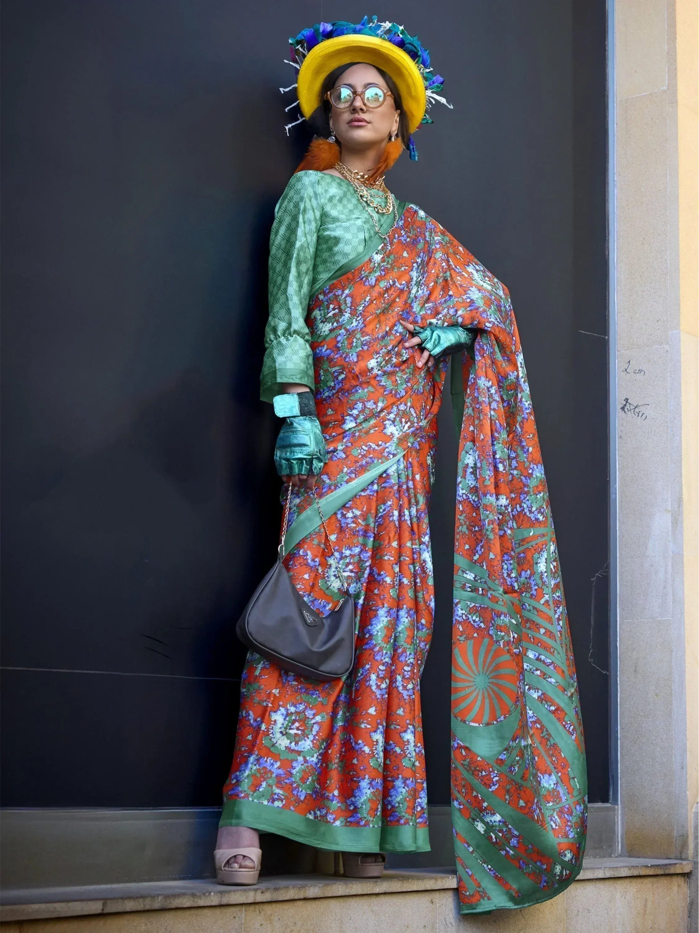 Woman in vibrant orange floral saree with green blouse, yellow feathered hat, and stylish accessories