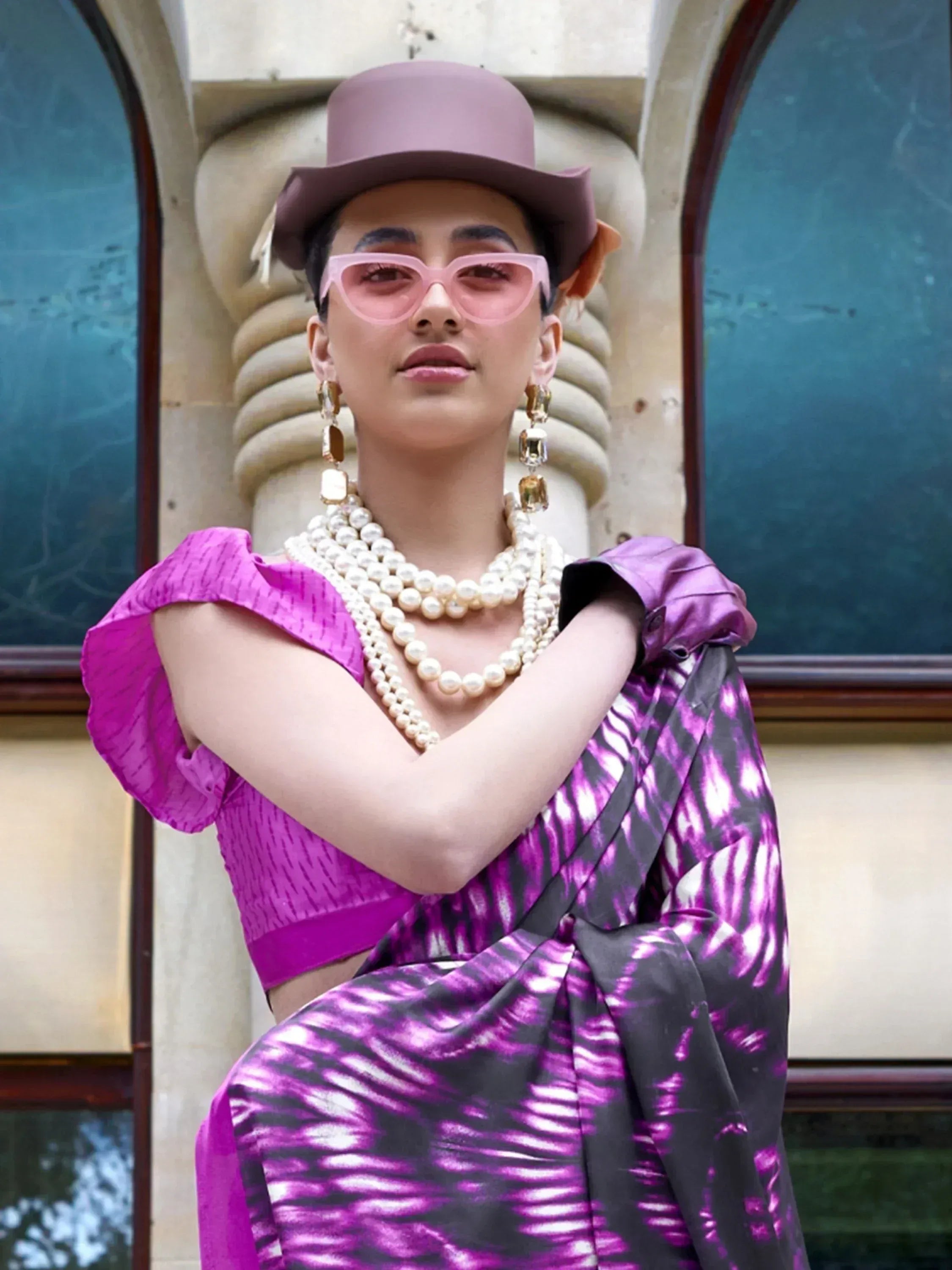 Woman in purple tie-dye saree with puff sleeves, pearl necklace, pink sunglasses, and hat