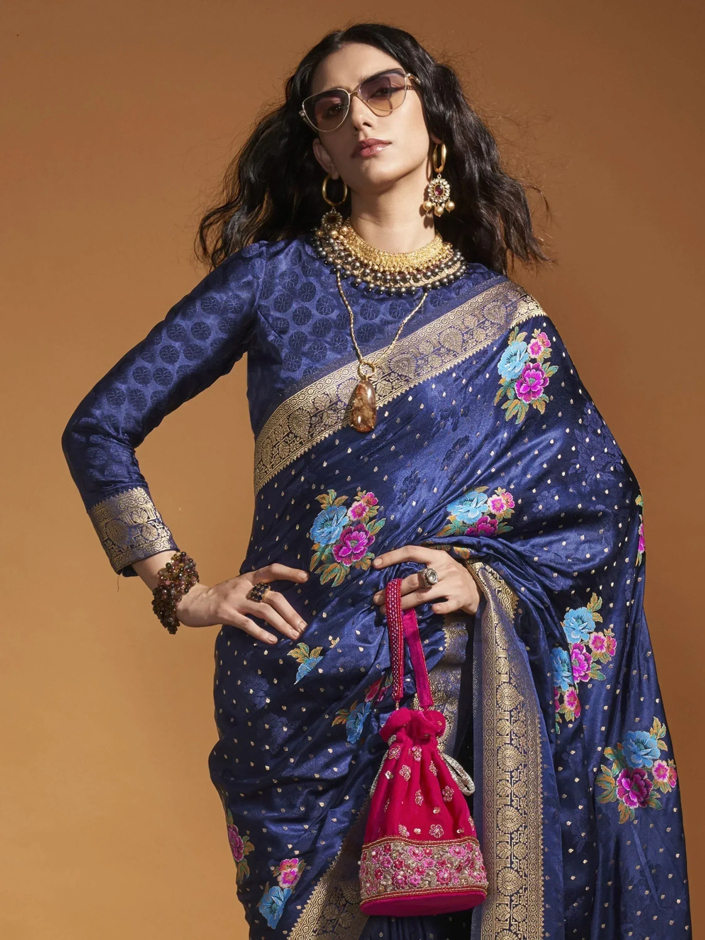 Woman in royal blue floral embroidered saree with gold border, paired with statement jewelry and pink potli bag