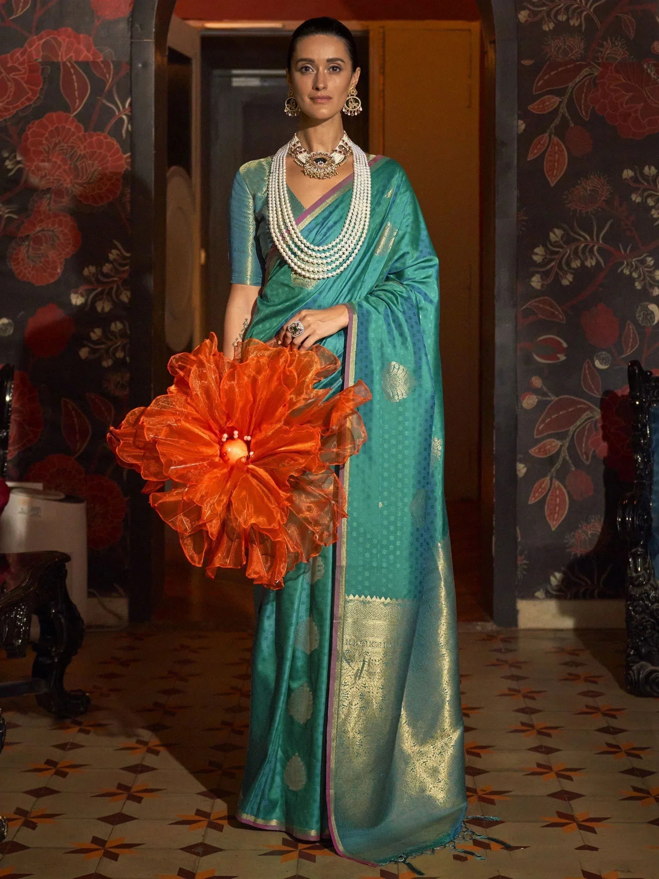 Teal silk saree with gold motifs, pearl necklace, and orange floral accessory for Indian ethnic wear