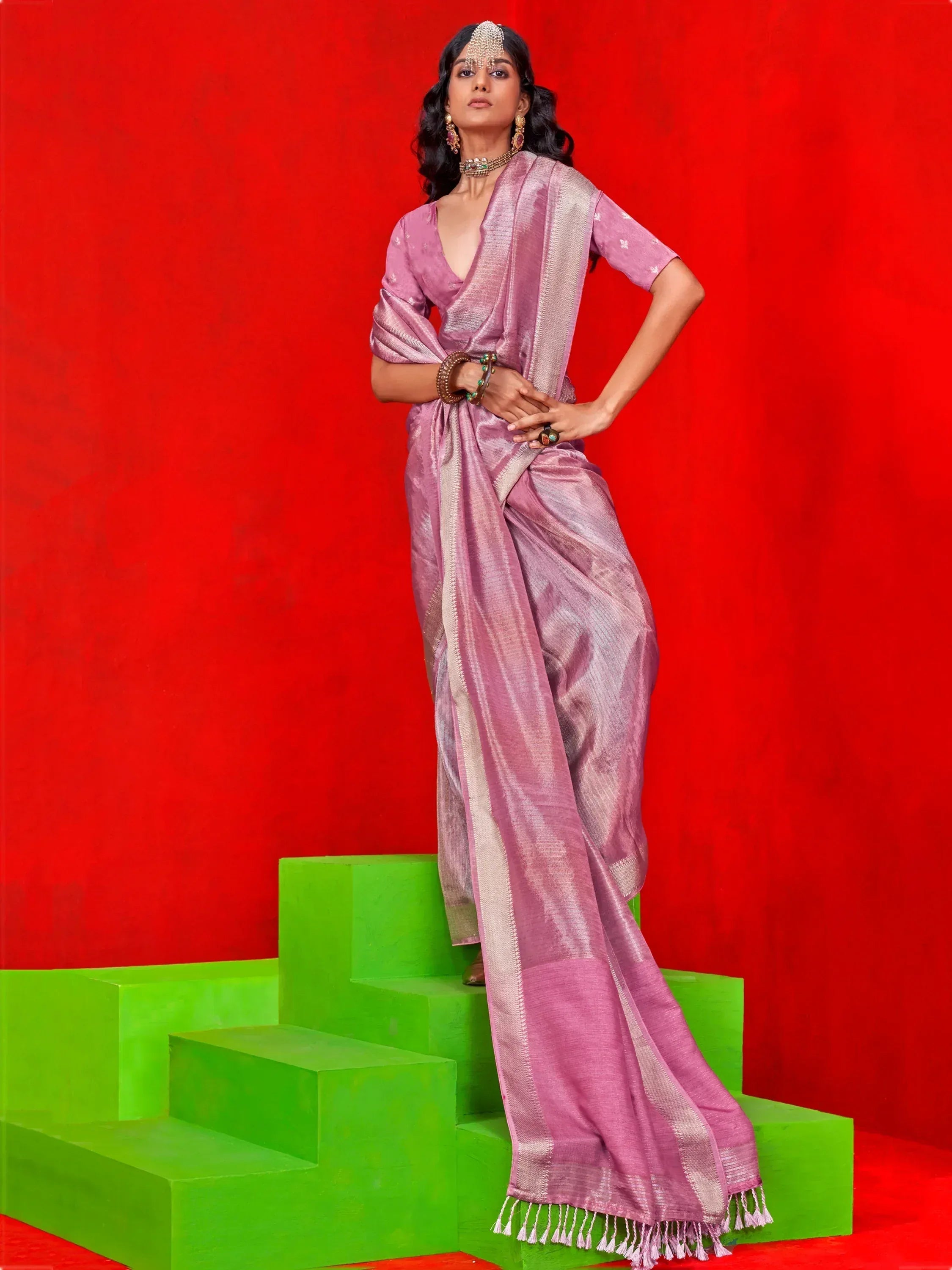 Woman in pink ethnic saree with gold border, standing on green steps against red background
