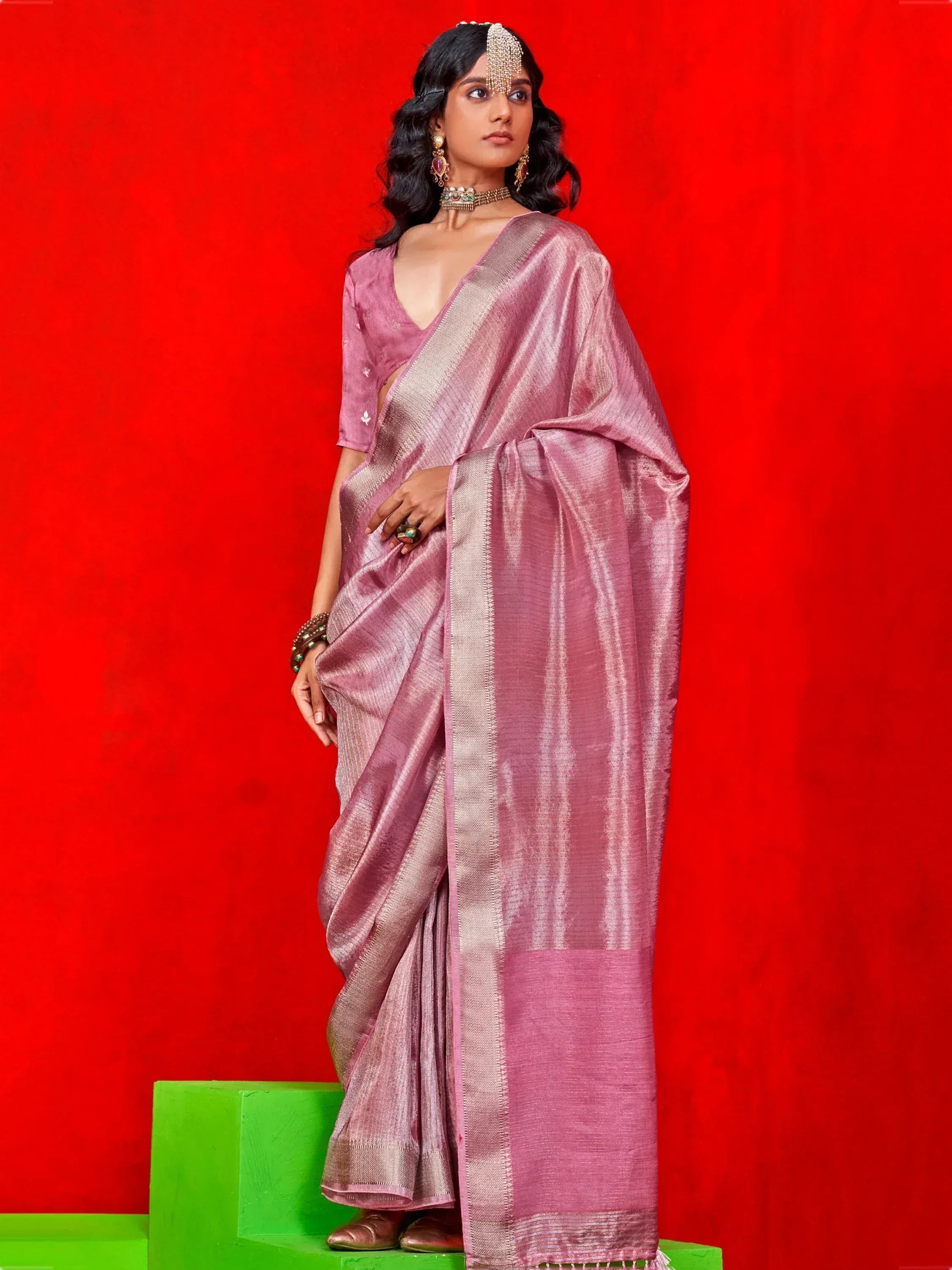 Pink silk saree with gold border, traditional Indian ethnic wear for women, styled with jewelry against red backdrop