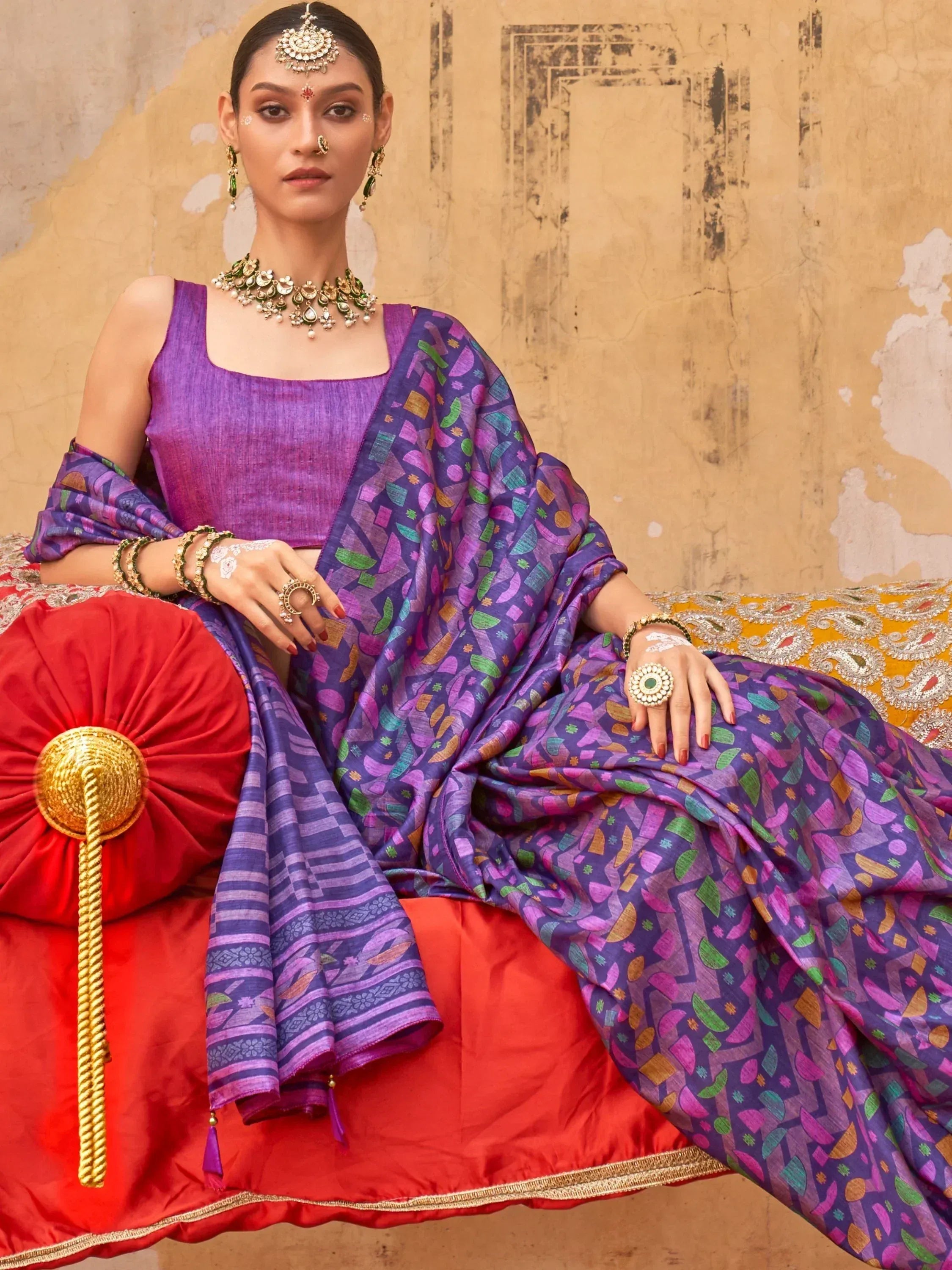 Woman in purple geometric print saree with matching blouse, traditional jewelry, seated on red cushion against rustic wall