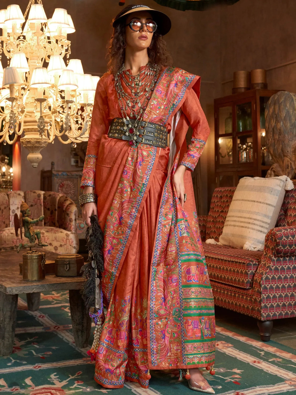 Woman in vibrant orange ethnic saree with intricate paisley and elephant prints, styled with belt and sunglasses