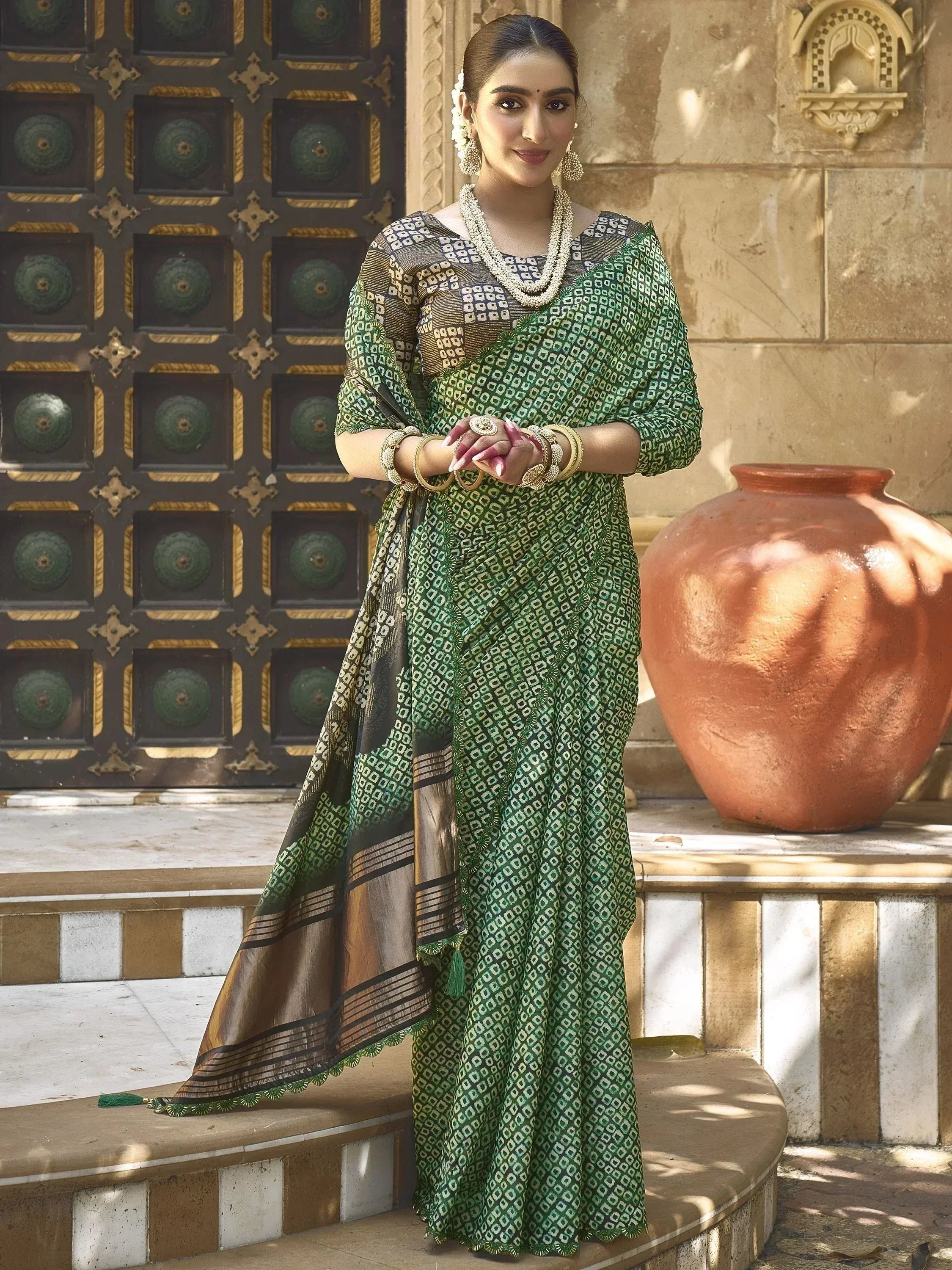 Woman in green and gold printed saree with traditional jewelry, standing by ornate door and clay pot
